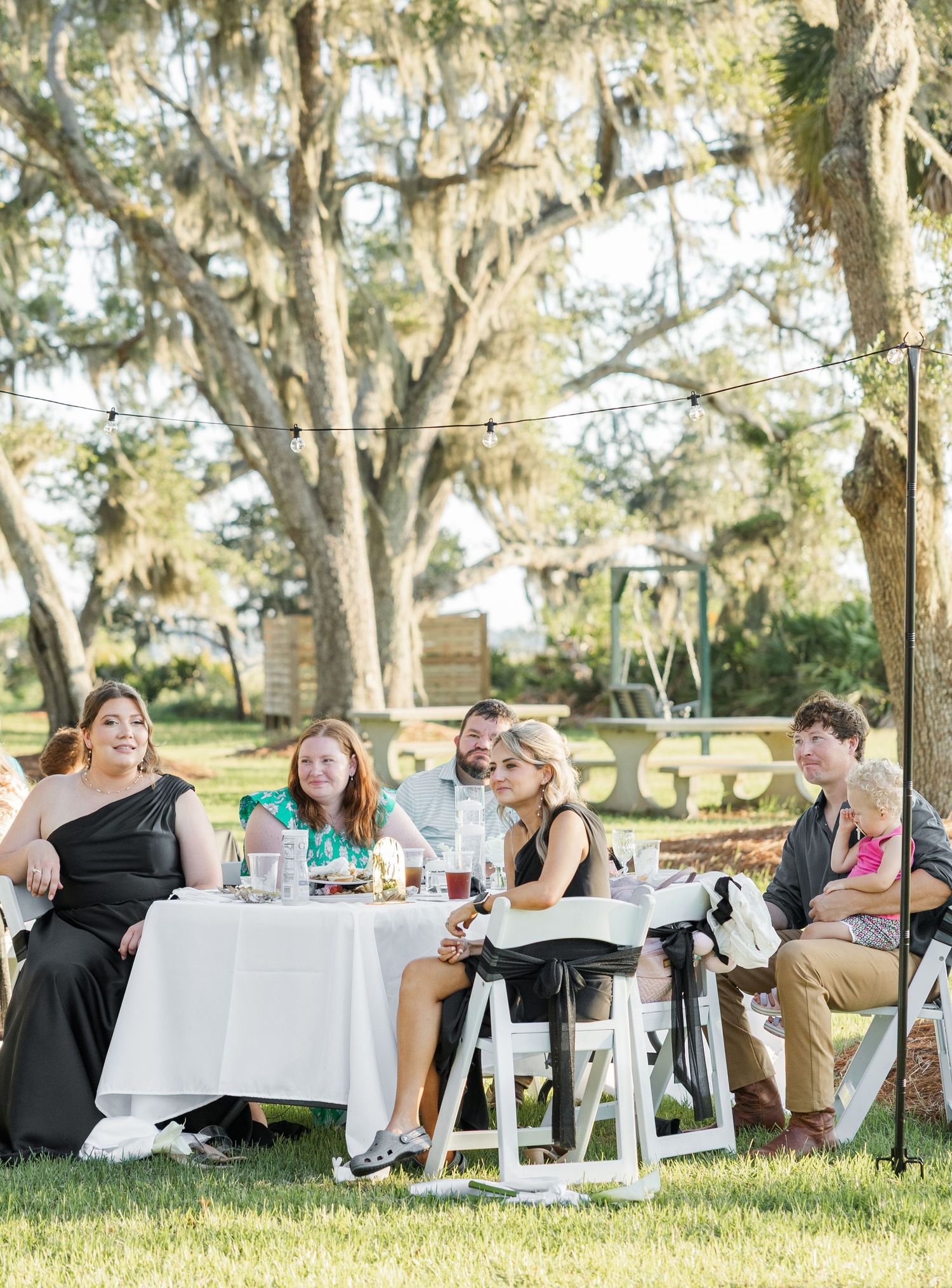 Wedding guests enjoy dinner at white-draped tables under Spanish moss-covered trees outdoors.