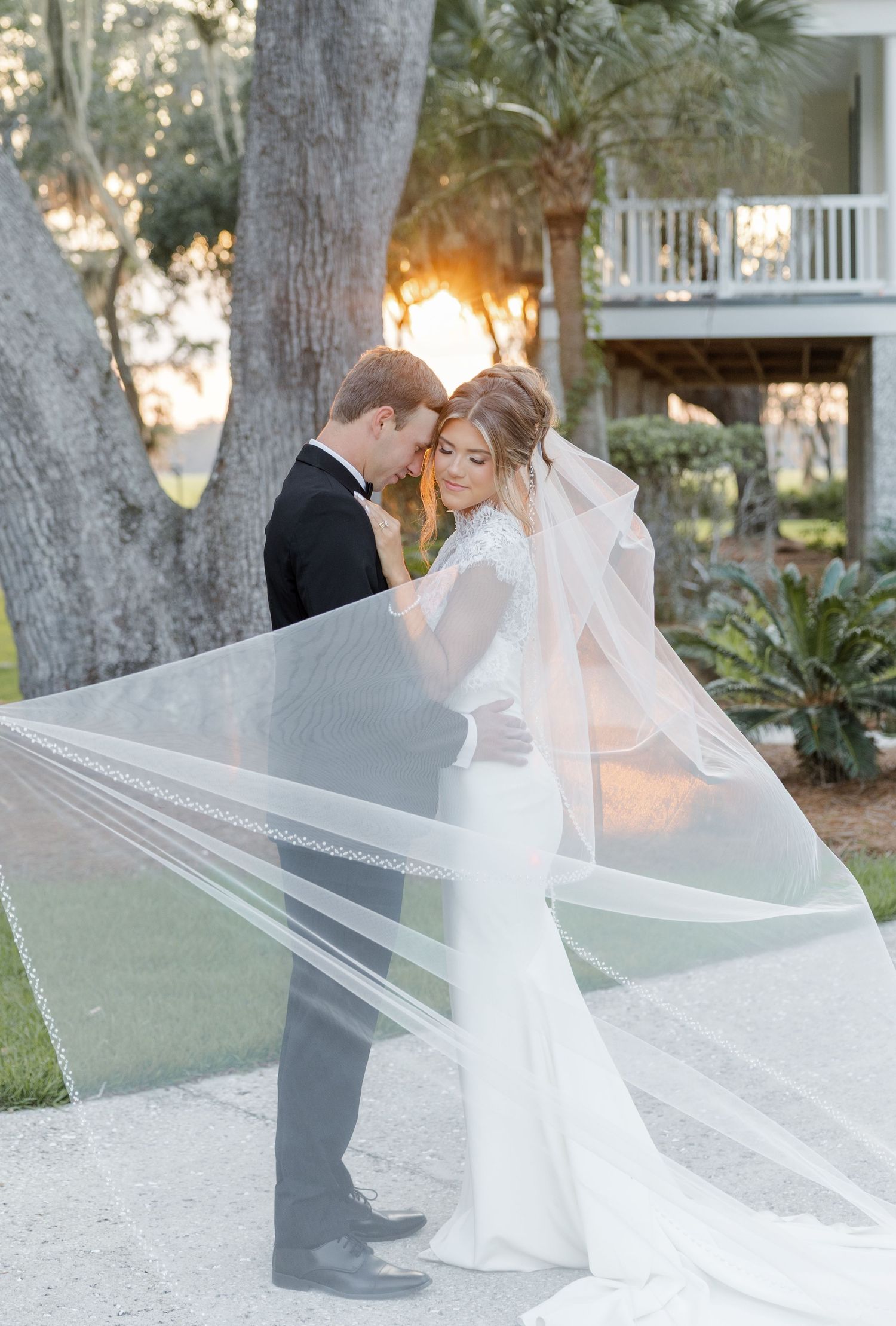 Bride and groom share romantic moment as cathedral veil billows in golden sunset light outdoors.