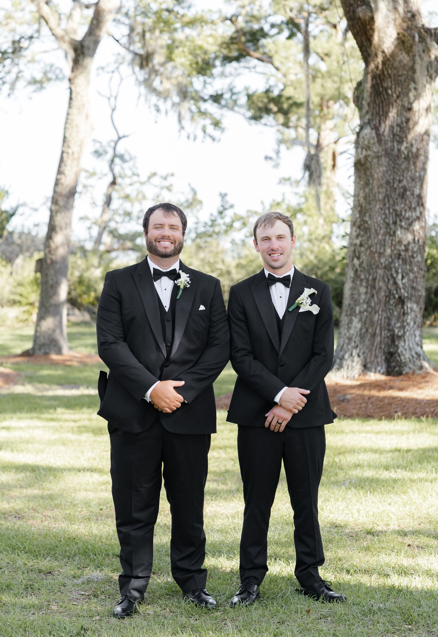 Two groomsmen in black tuxedos with bow ties and boutonnieres posing outdoors under oak trees.