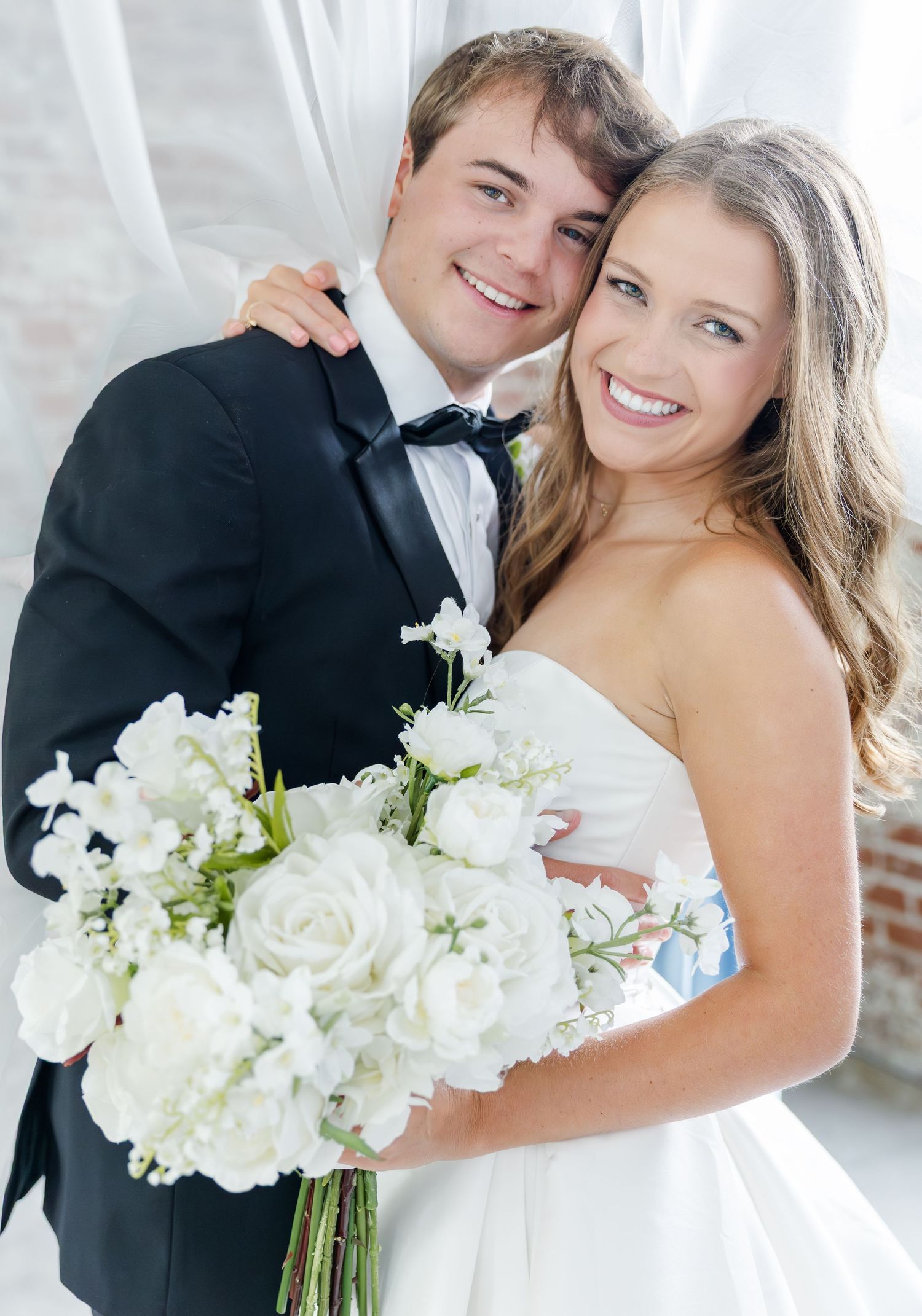 Smiling bride and groom pose together, bride holding white rose bouquet, groom in black tuxedo with bow tie.
