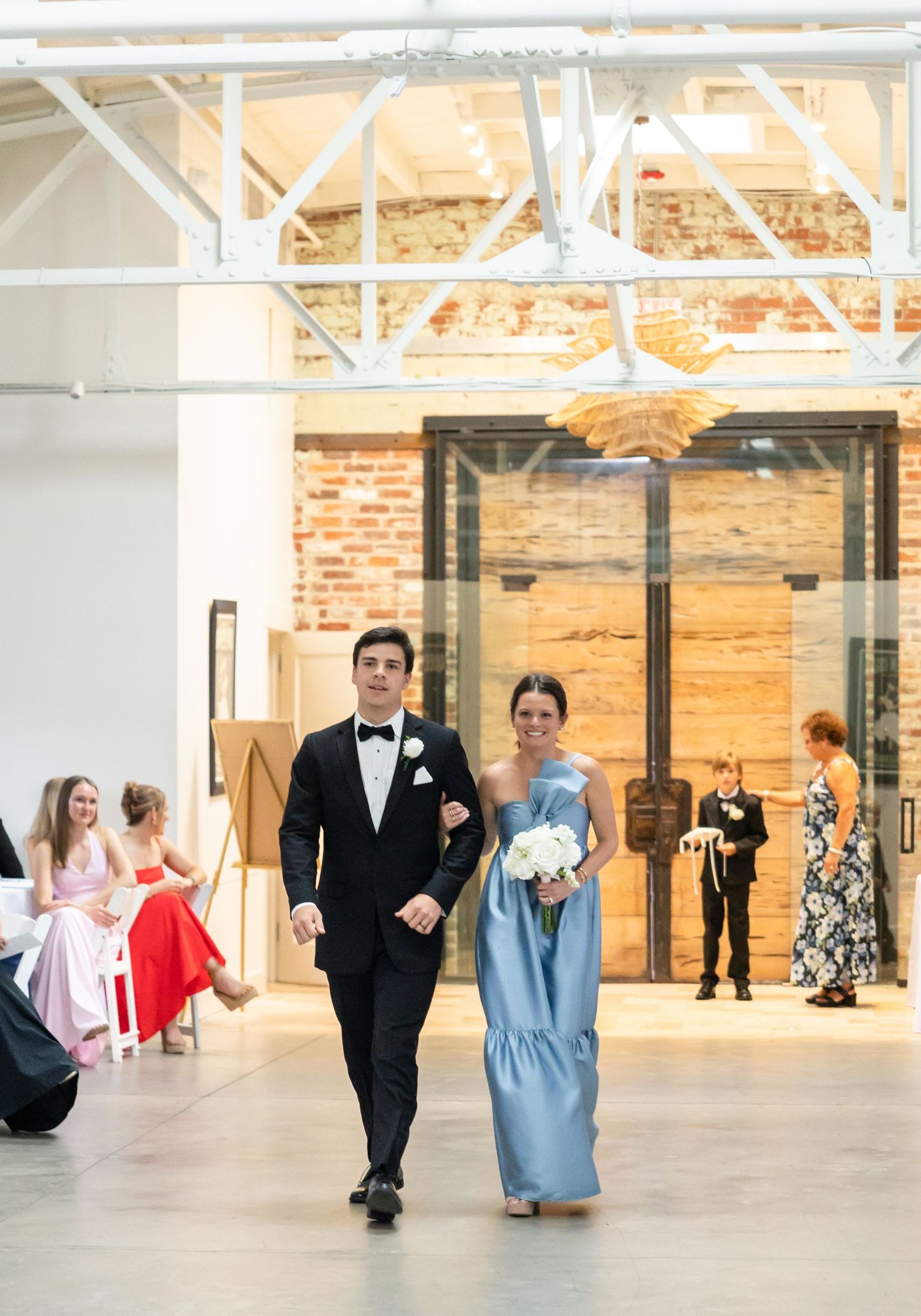 Groomsman escorts bridesmaid in blue gown down the aisle at modern industrial wedding venue with exposed brick walls.