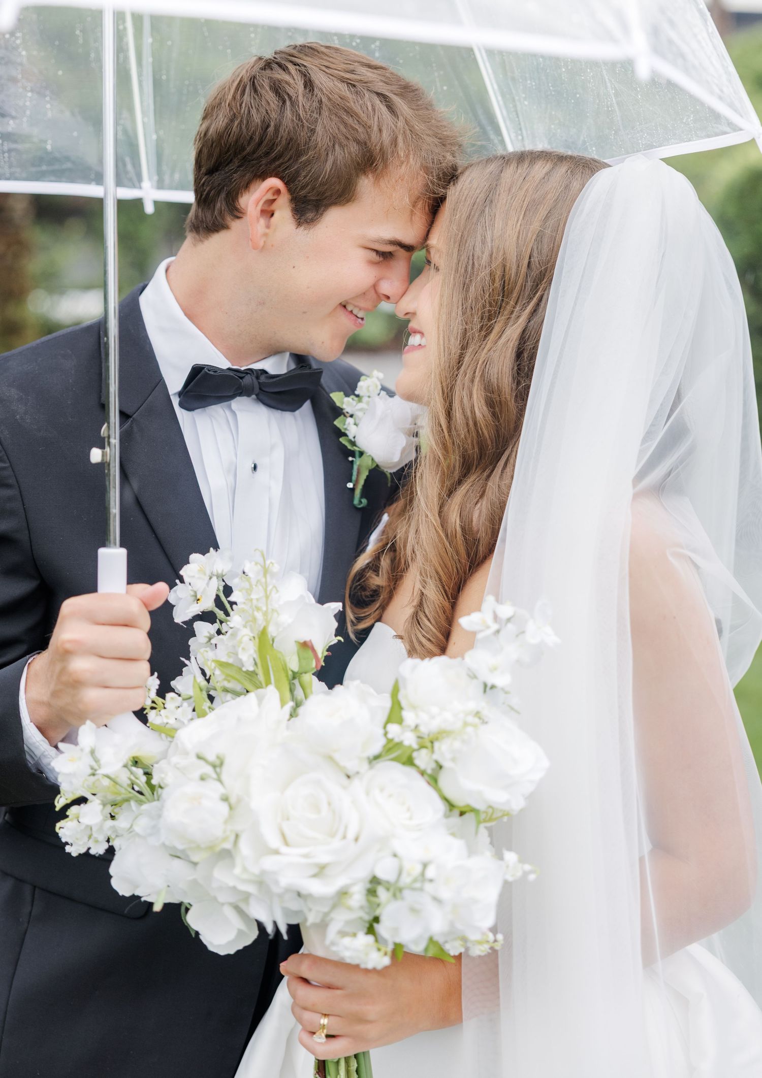 Bride and groom share a romantic moment under a clear umbrella, bride holding white rose bouquet.