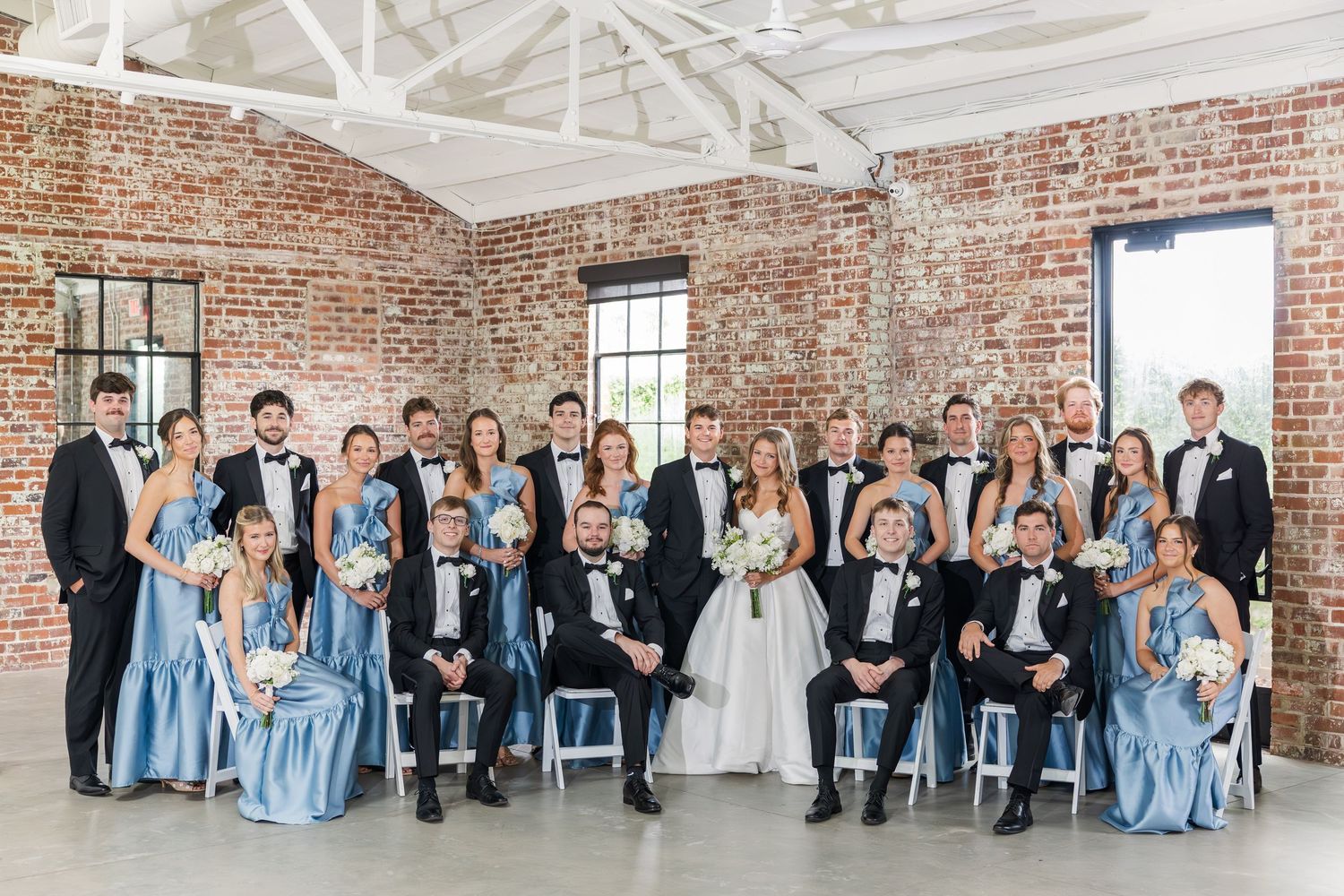 Large wedding party posing in blue bridesmaid dresses and black tuxedos inside a brick venue.