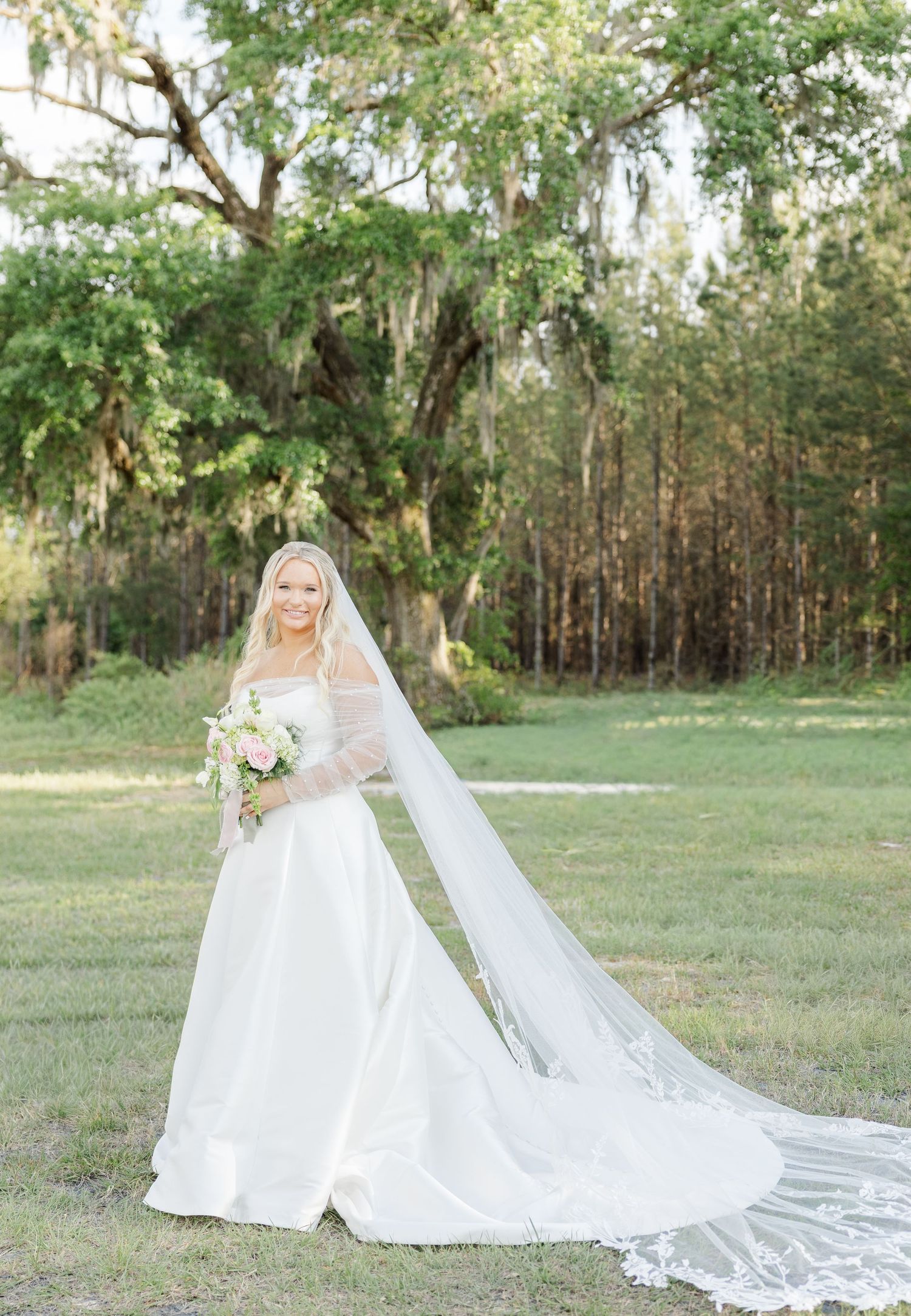 Bride in elegant white ball gown with long lace-trimmed cathedral veil holding pink bouquet outdoors on green lawn.