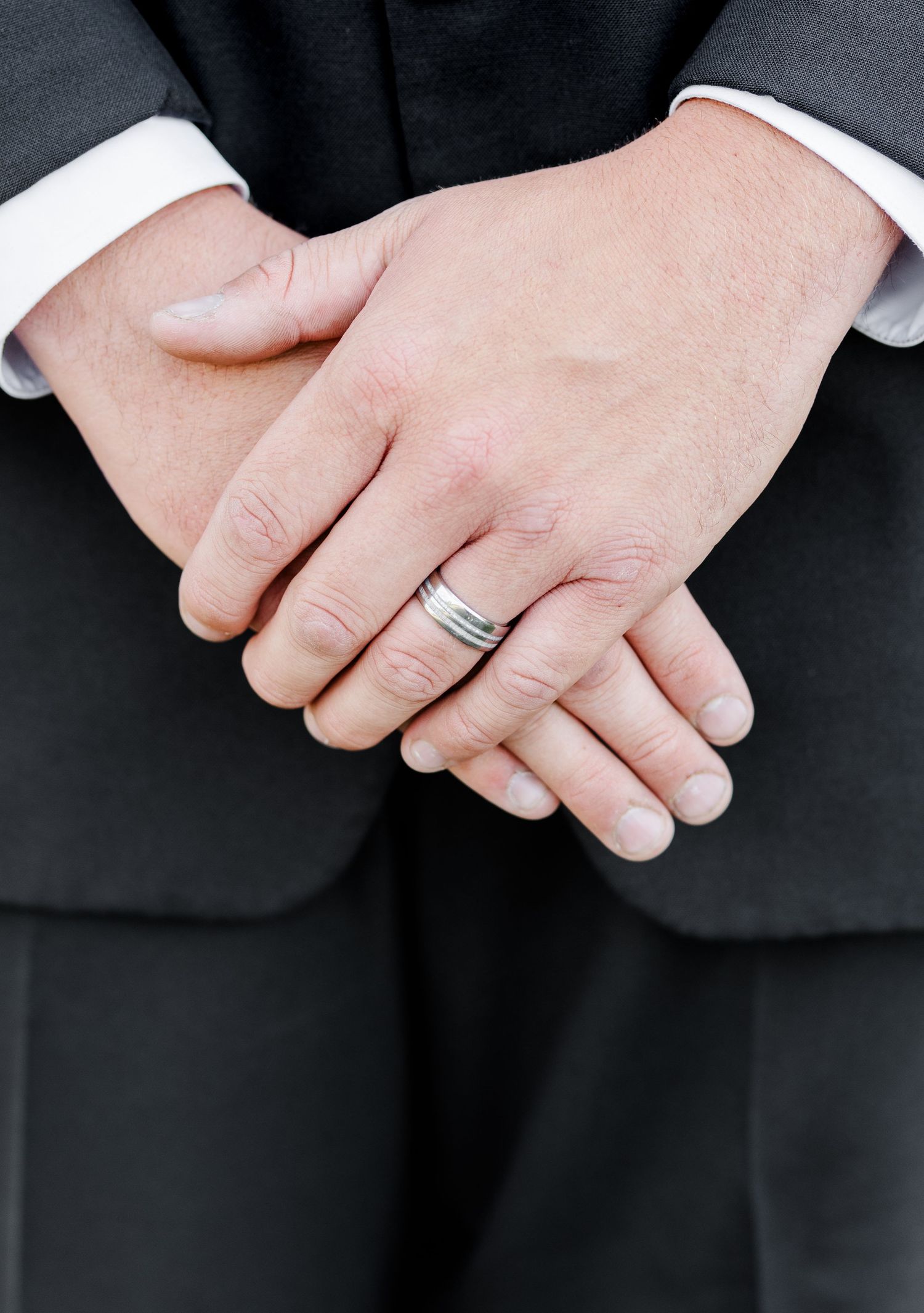 Close-up of groom's hands clasped together wearing a silver wedding band against a dark suit.