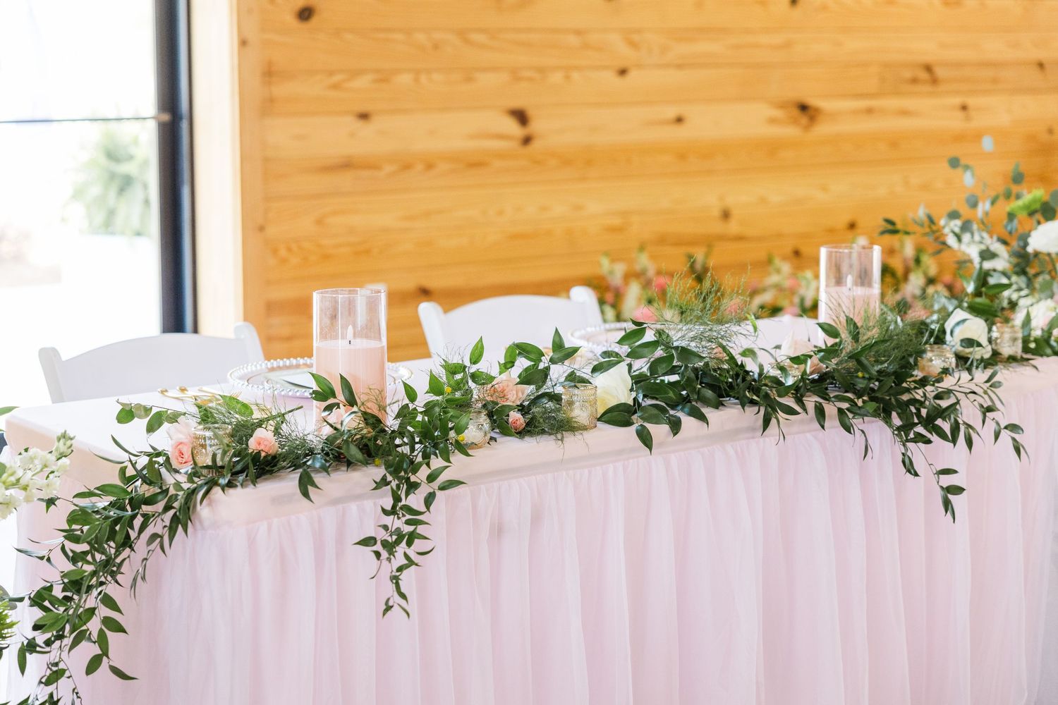 Elegant wedding head table with pink tulle skirt, lush greenery garland, white flowers, and candles in a wood-paneled venue.