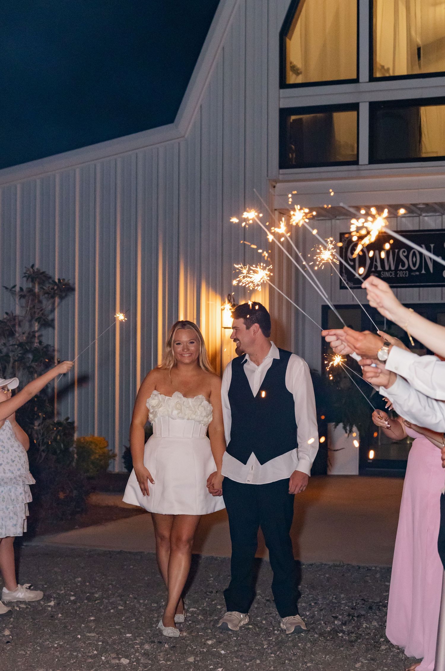 Newlyweds walk through sparkler send-off exit at night outside barn wedding venue.