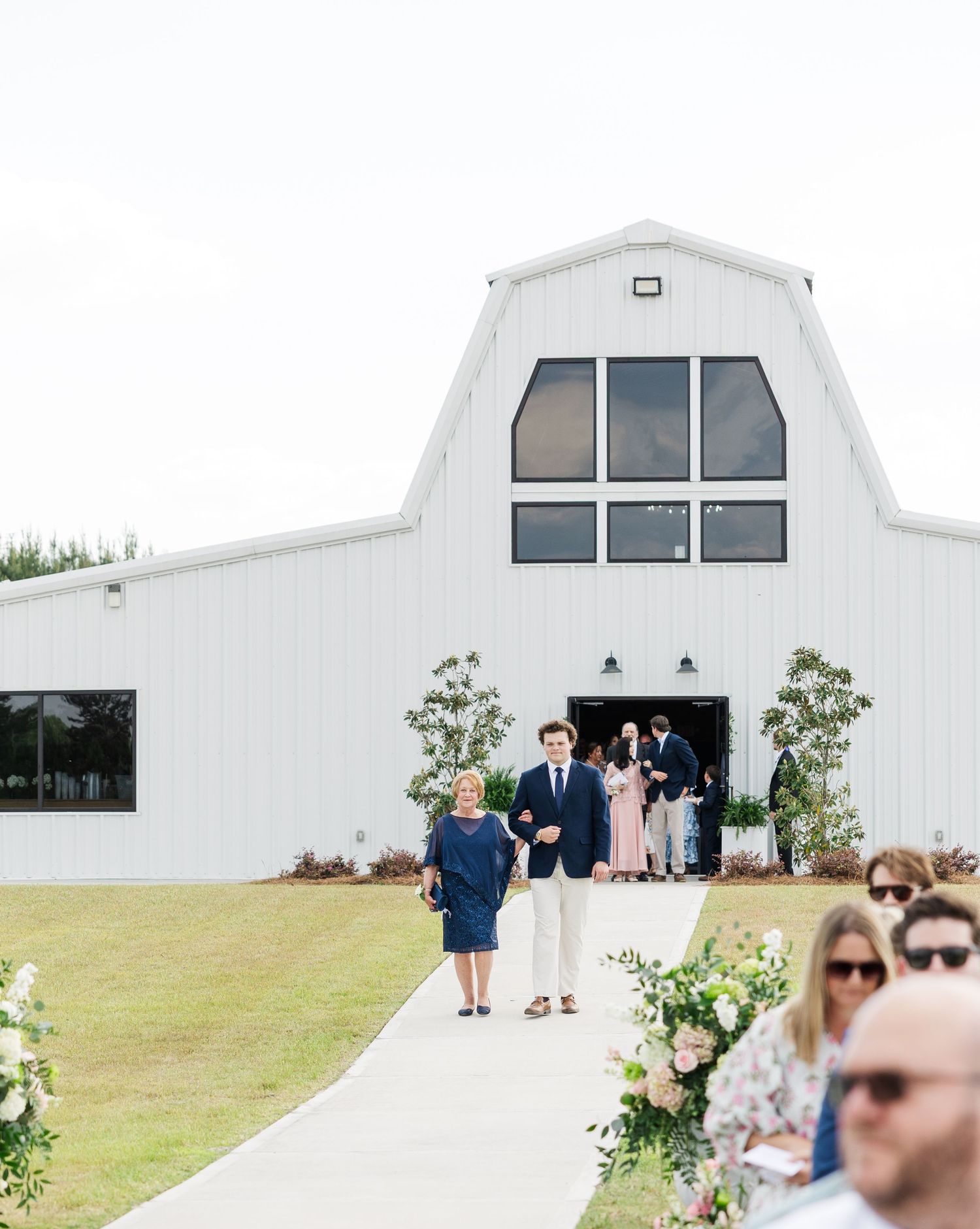 Outdoor wedding ceremony at white barn venue with guests seated along floral-lined aisle.