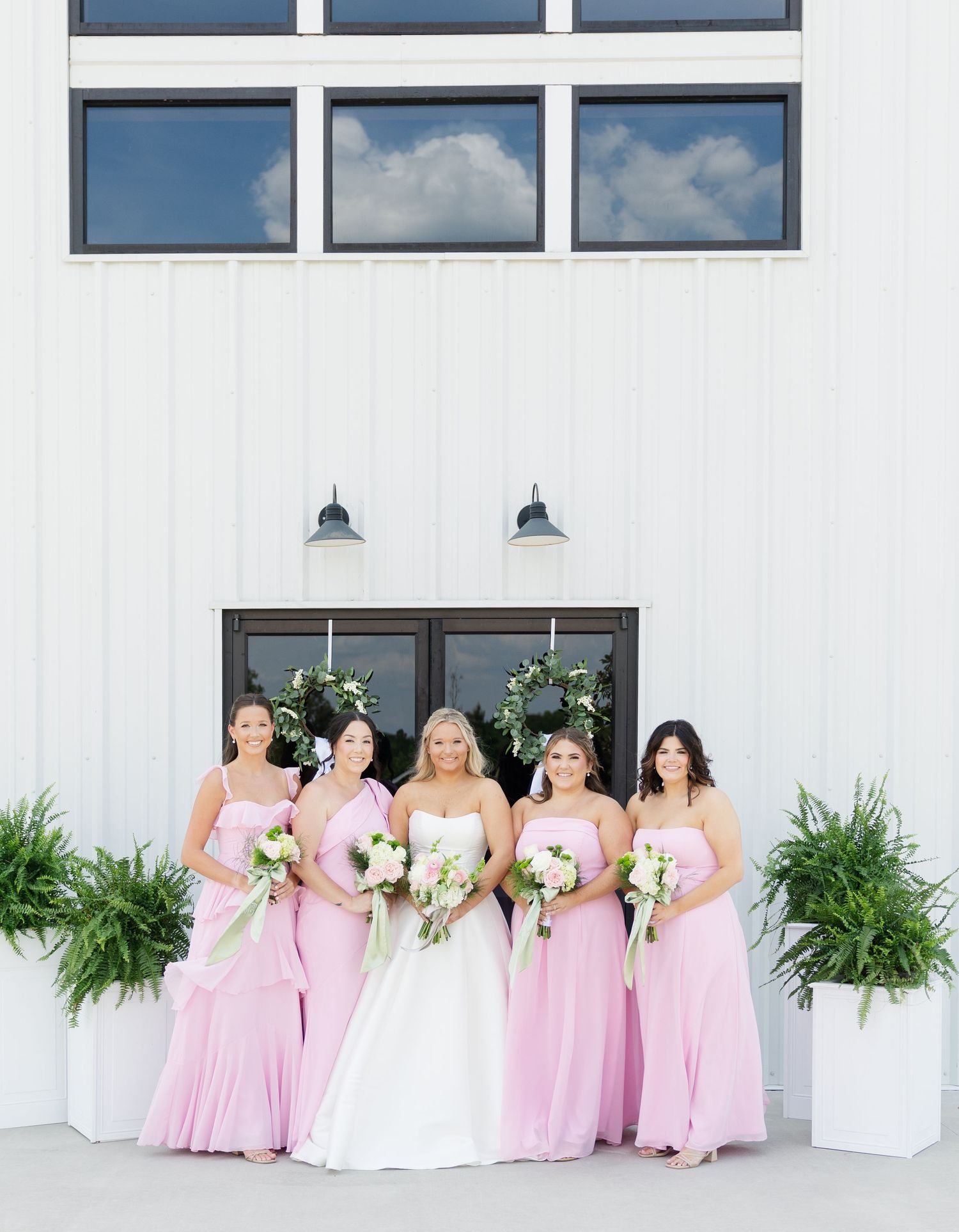 Bride in white gown poses with four bridesmaids in pink dresses holding bouquets outside a white barn venue.