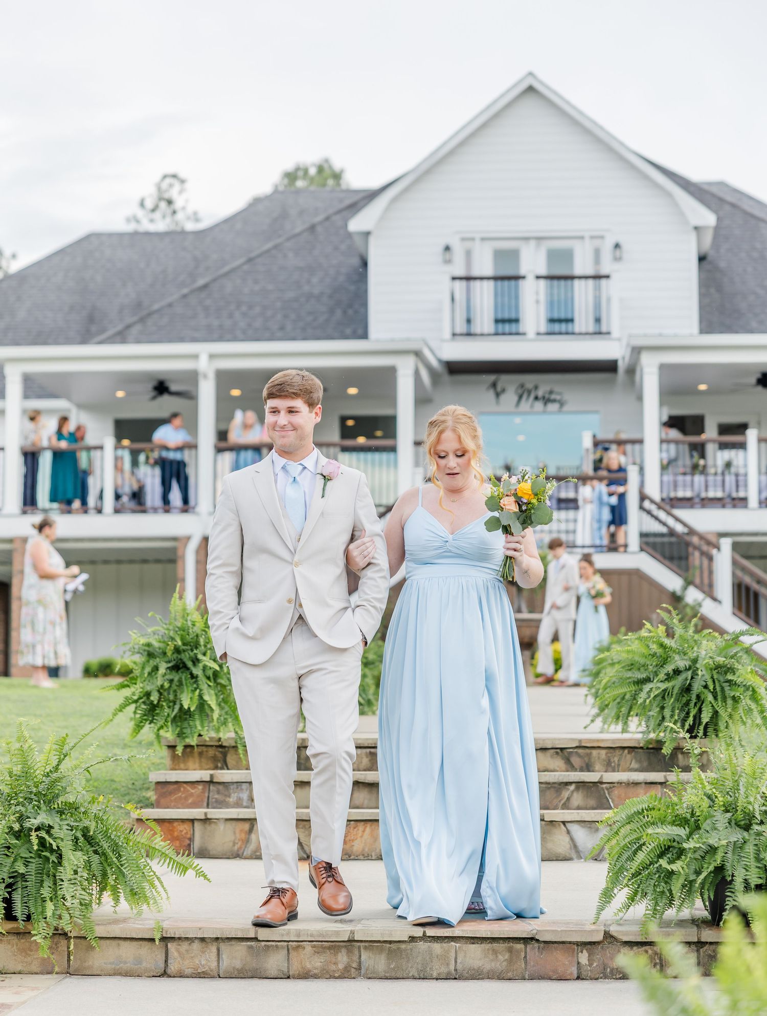 Groomsman escorts bridesmaid in blue gown down steps at outdoor wedding ceremony venue.