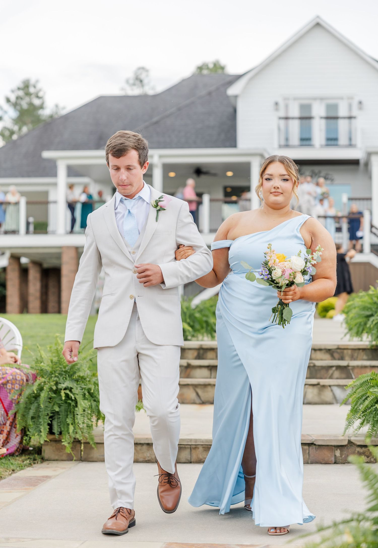 Groomsman in beige suit escorts bridesmaid in light blue gown down outdoor wedding aisle.