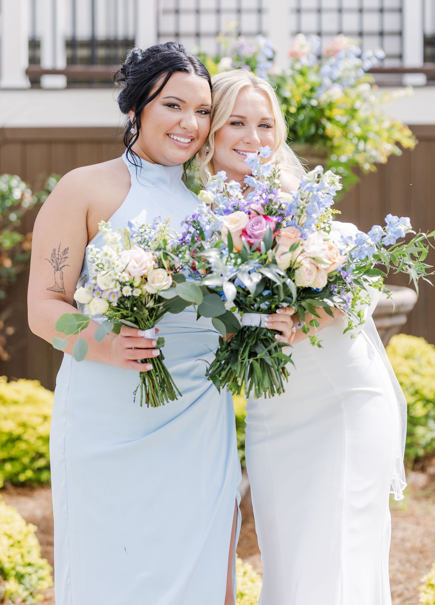 Bride and bridesmaid in light blue halter gown smile together holding colorful floral bouquets outdoors.