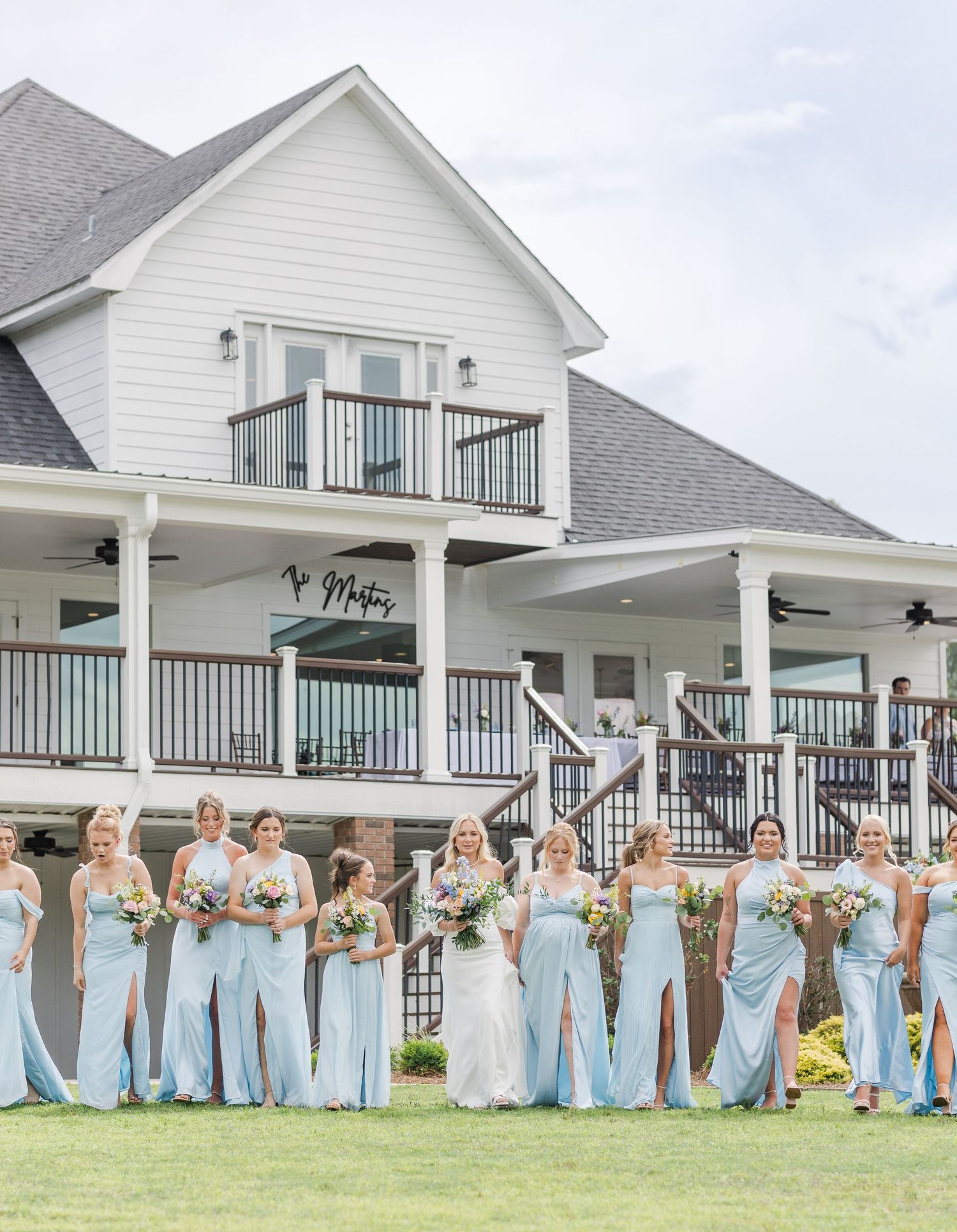 Bride poses with large bridal party in light blue gowns in front of white farmhouse wedding venue.