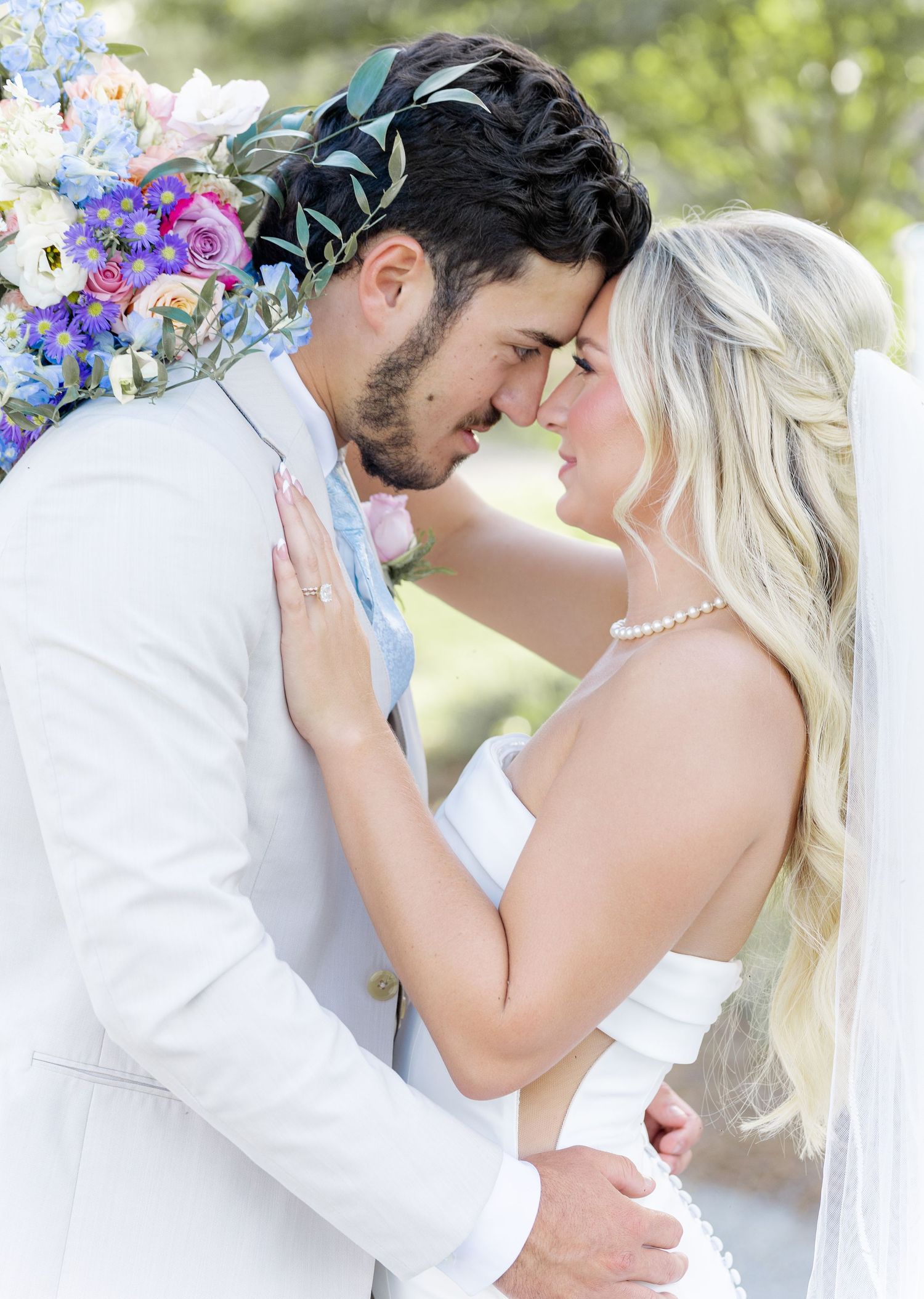 Bride and groom in white attire share a romantic forehead touch surrounded by colorful wedding bouquet outdoors.