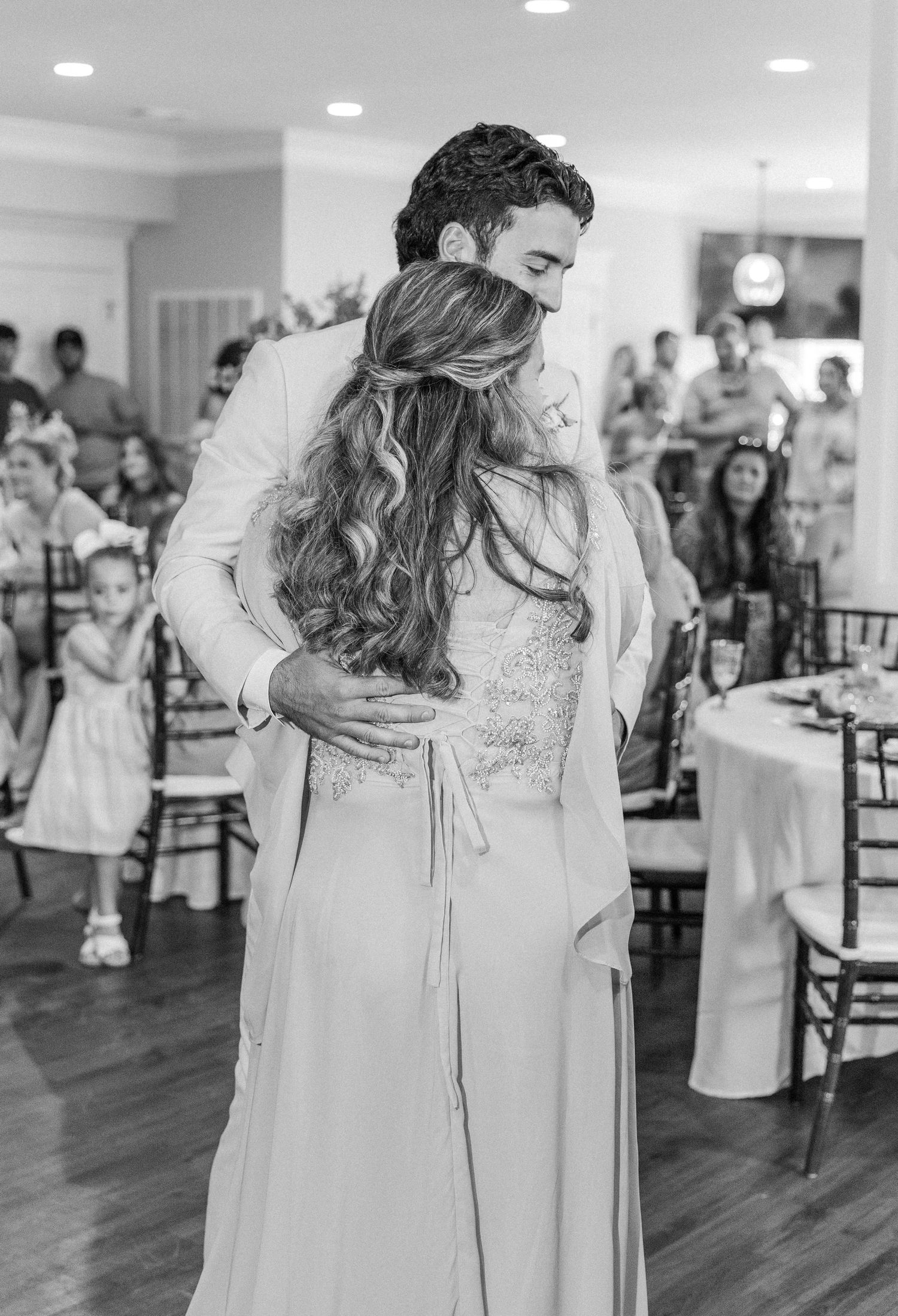 Bride and groom share first dance embrace in elegant white reception hall surrounded by guests.