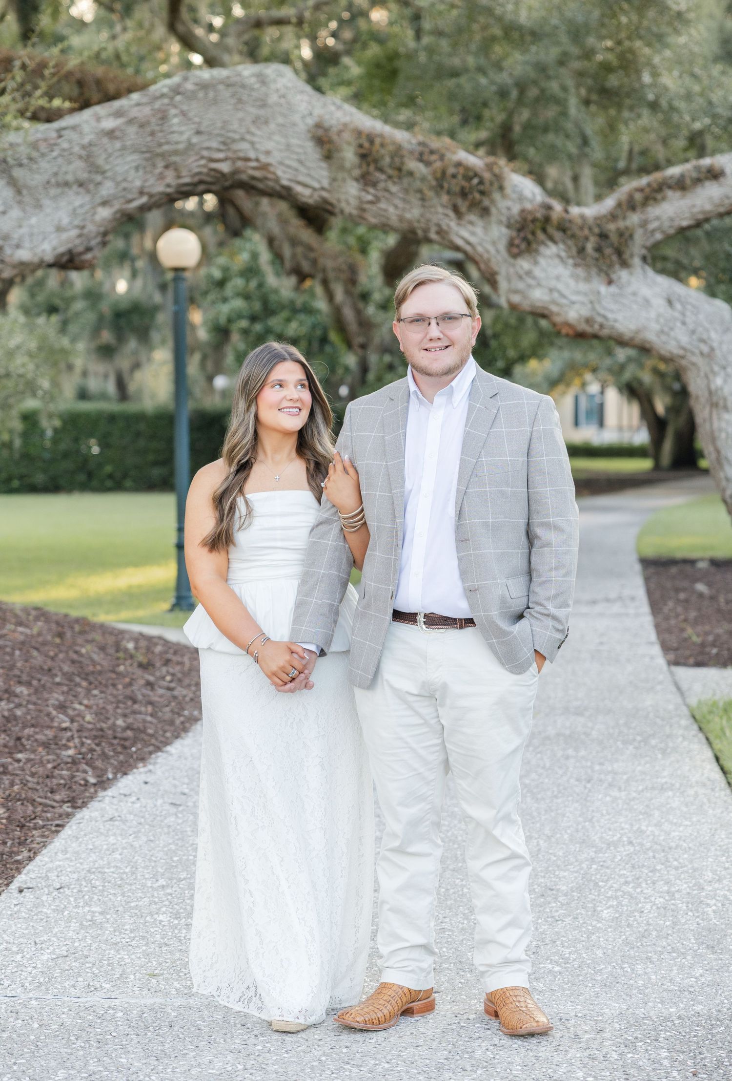 A couple in white and gray outfits pose on a tree-lined pathway during an outdoor engagement photo session.