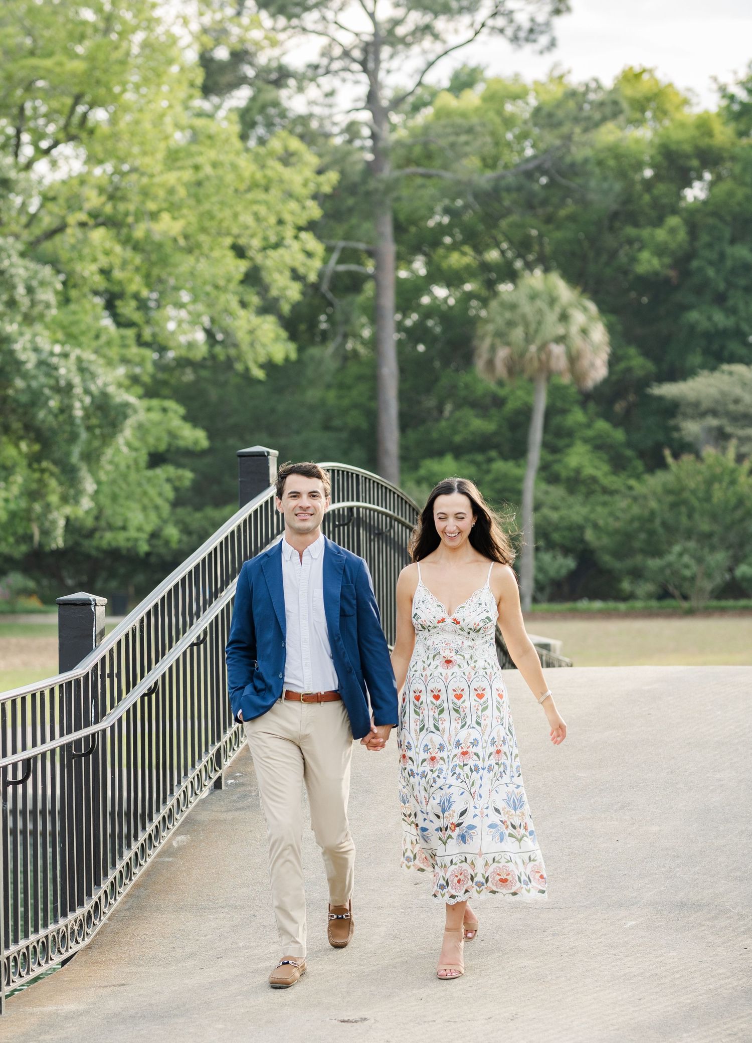 Smiling couple walking hand-in-hand across a bridge surrounded by lush green trees during an engagement session.