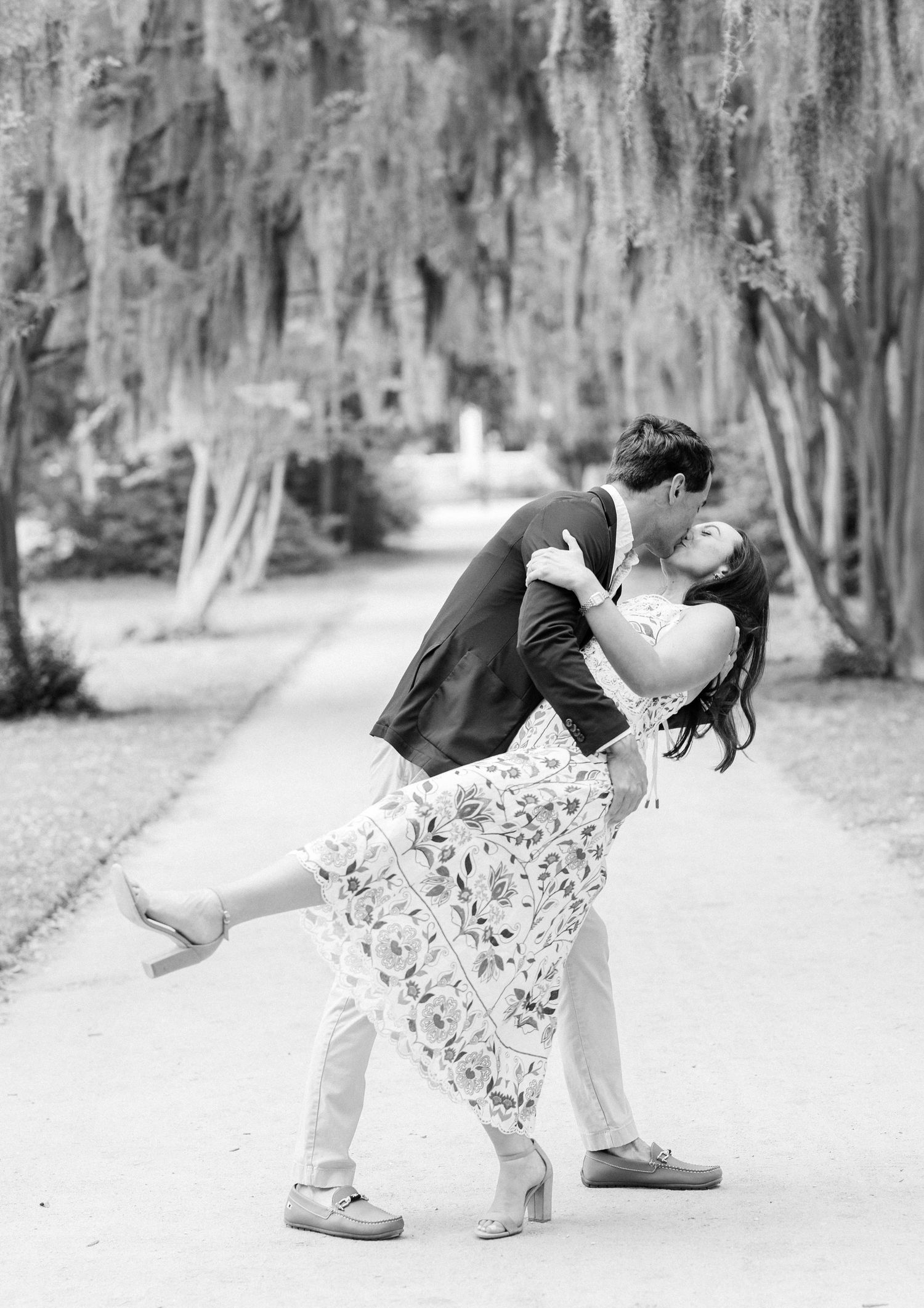 Black and white photo of a couple sharing a romantic dip kiss on a tree-lined path draped with Spanish moss.