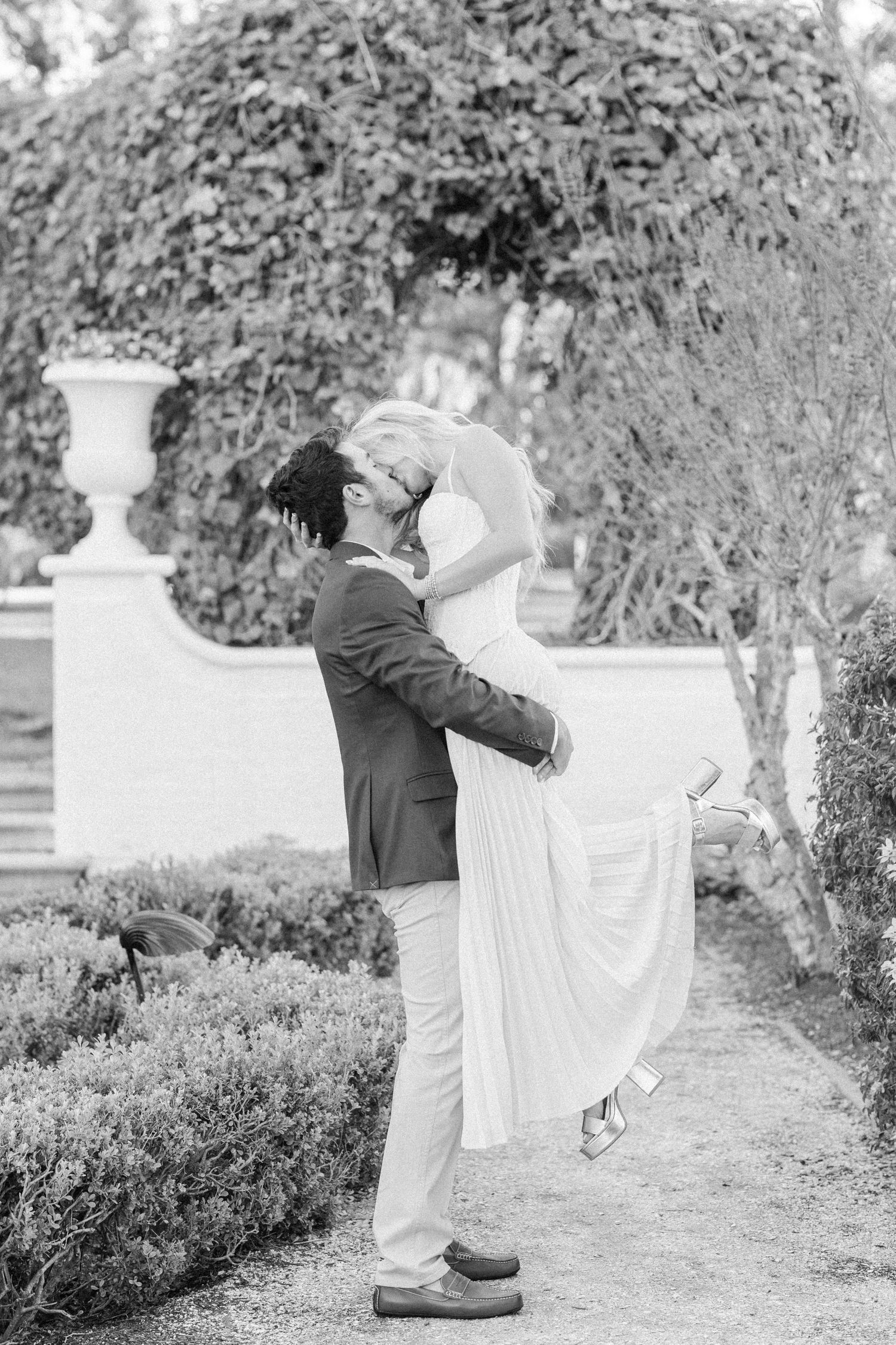 A romantic black and white photo of a couple embracing in a garden with decorative urns and lush foliage.