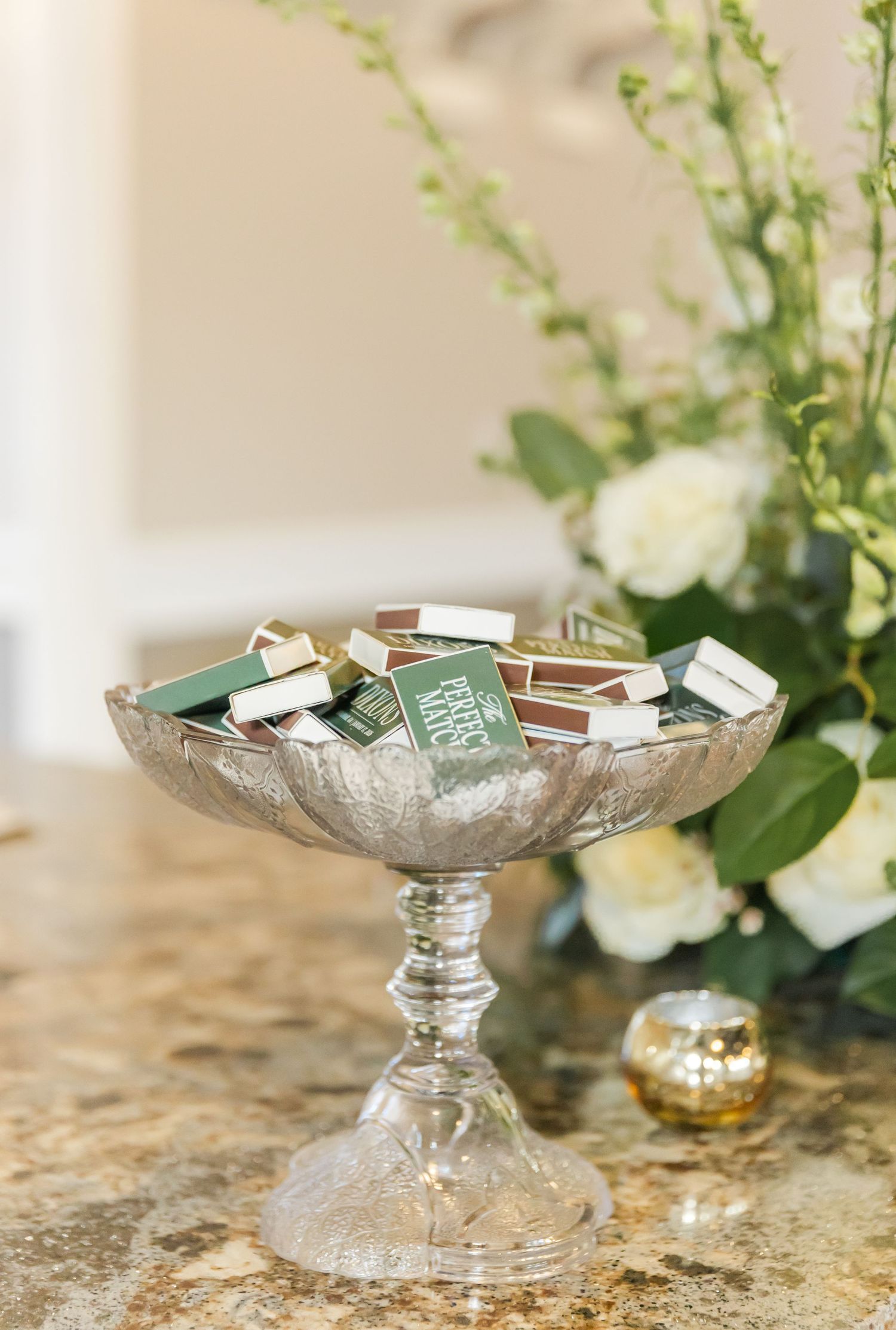 Elegant crystal pedestal dish displays place cards and wedding favors on a marble surface with white floral decor.