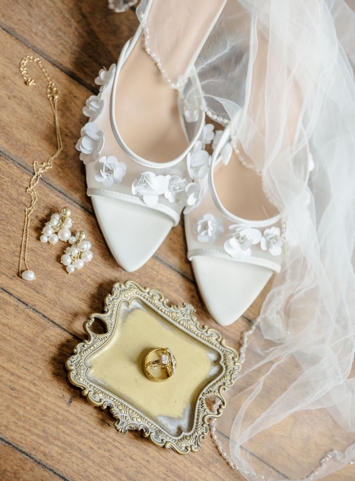 Elegant white bridal heels and veil arranged with delicate pearl jewelry and gold rings on rustic wooden surface.