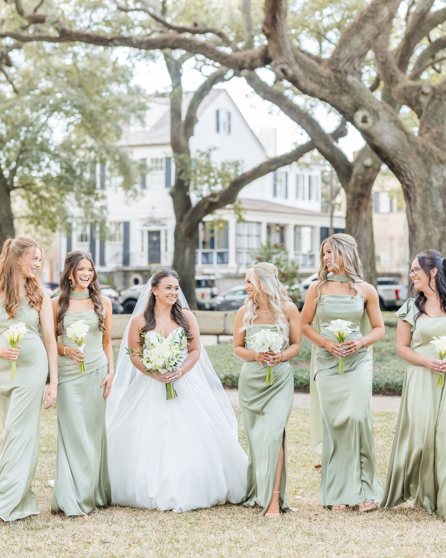 Wedding party in sage green bridesmaid dresses gathered under a large oak tree on a sunny day.