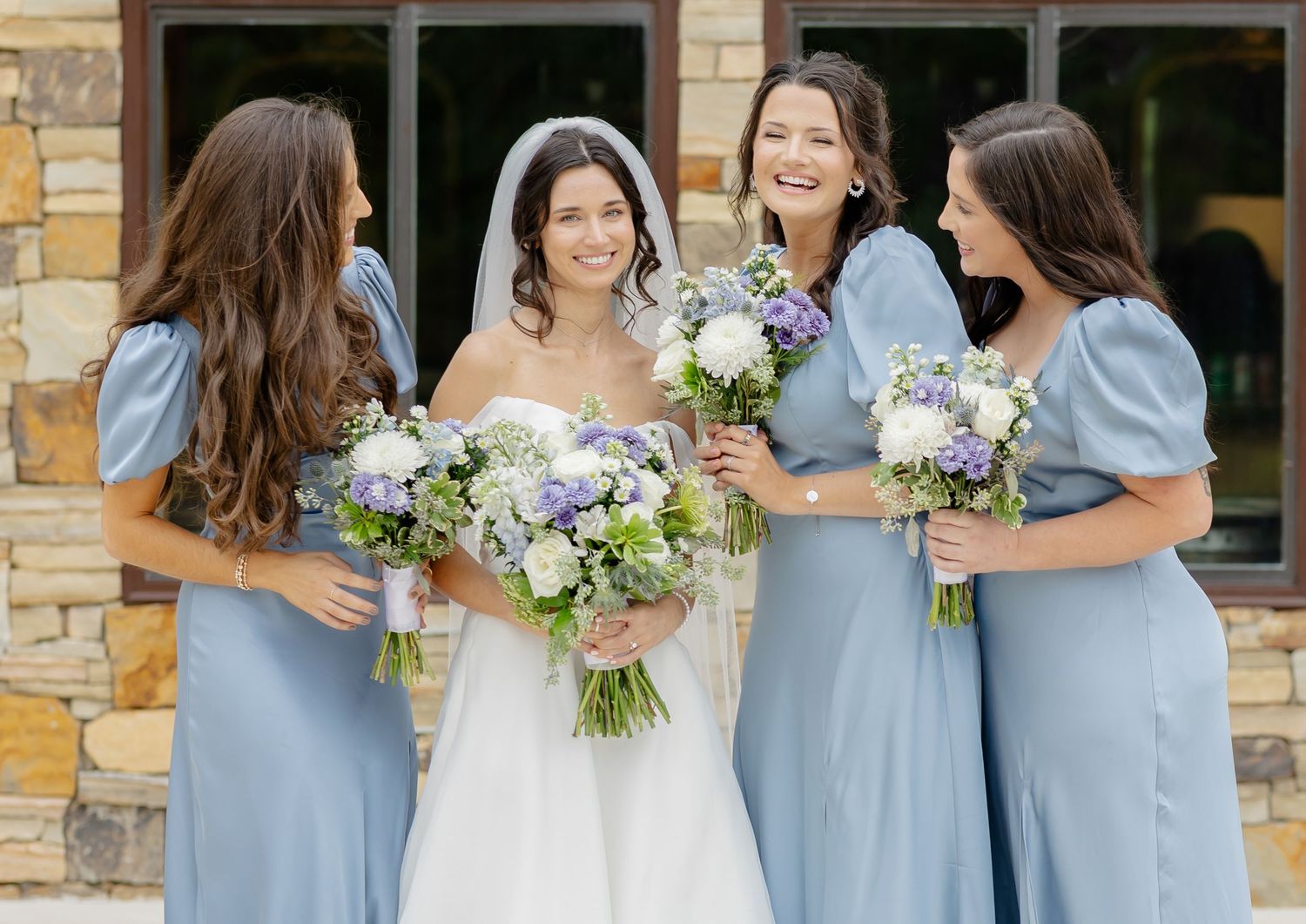 Bridesmaids in light blue dresses holding white and green bouquets stand together at an outdoor wedding venue.