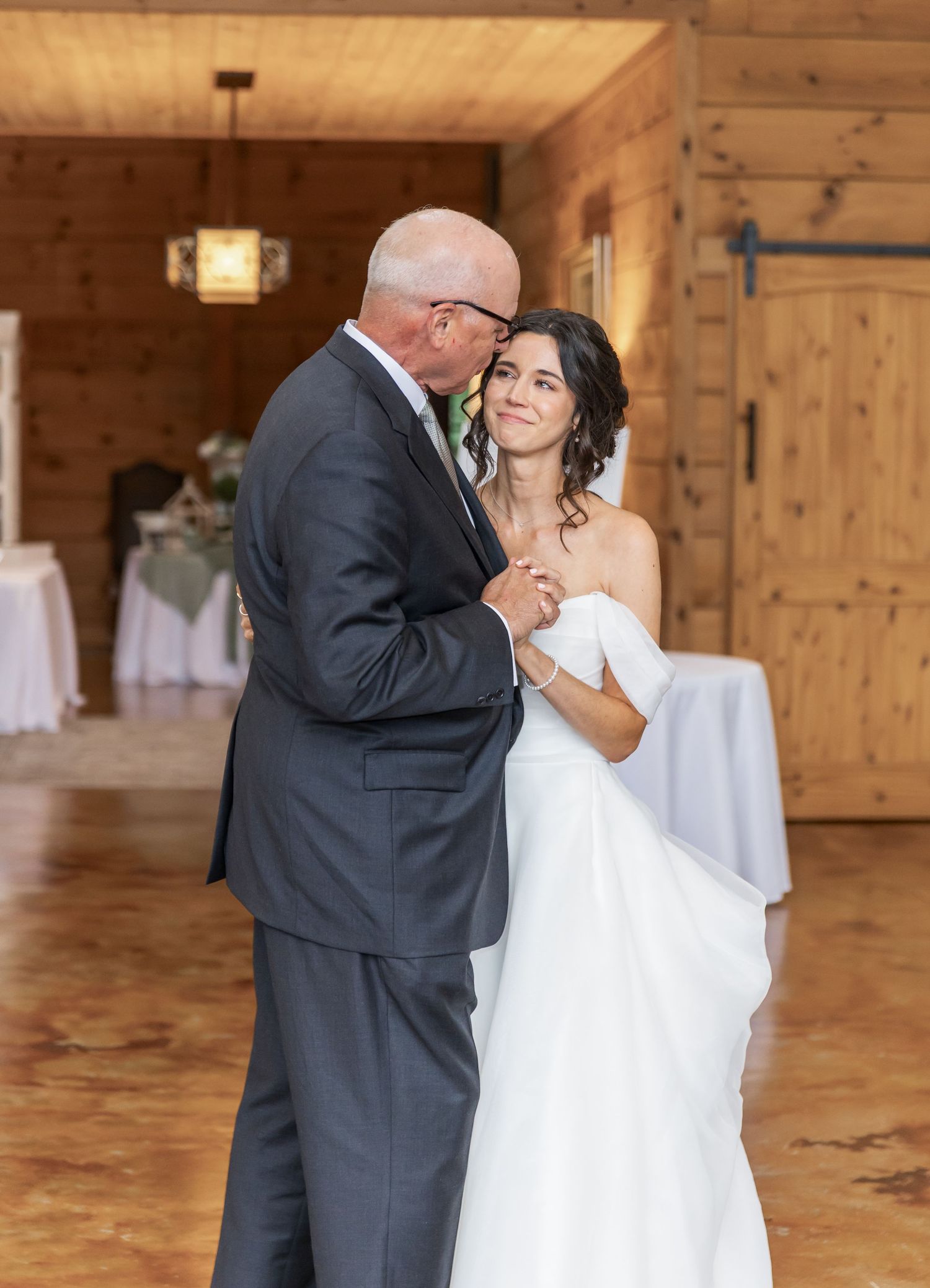 A tender dance moment captured in a rustic wood-paneled reception venue with white tablecloths.