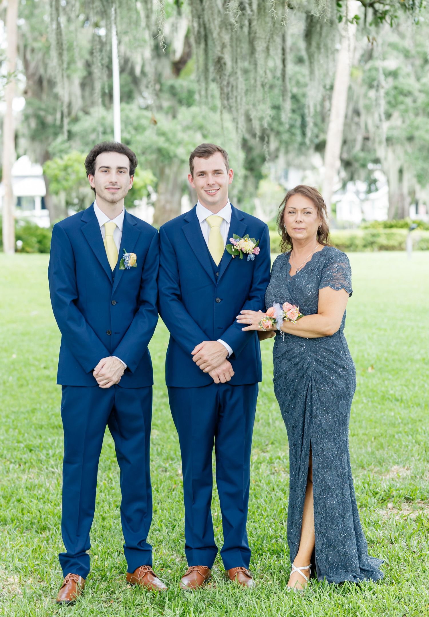 Three people in formal attire stand together on a grassy lawn with Spanish moss-draped trees in the background.