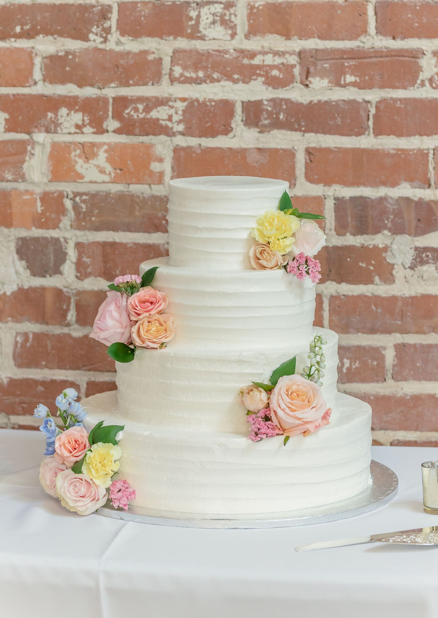 White tiered wedding cake decorated with delicate pastel roses and flowers against a rustic brick wall backdrop.