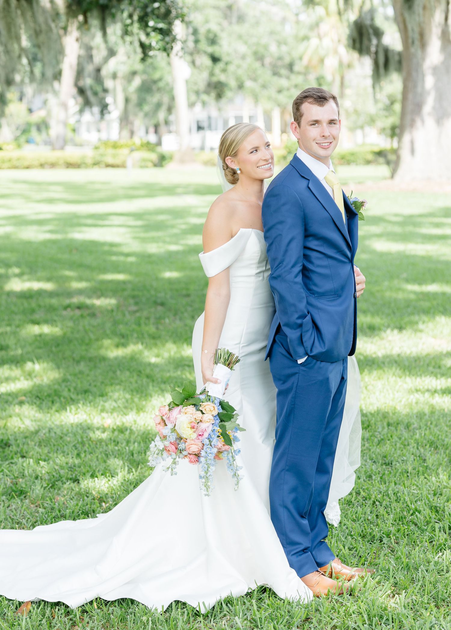A couple in wedding attire poses back-to-back on a lush green lawn with Spanish moss draped trees in the background.