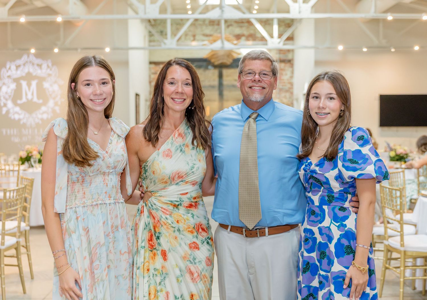 A group of four people in colorful summer attire pose together at an elegant indoor event venue with decorative lighting.