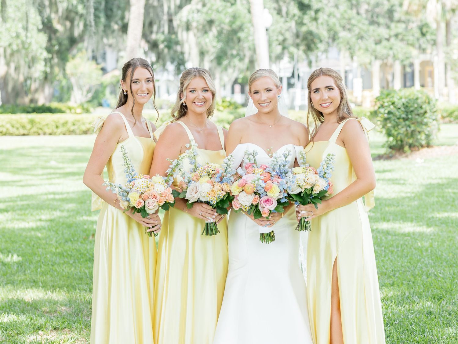 Bridesmaids in pastel yellow dresses hold colorful bouquets while posing outdoors in a garden setting.