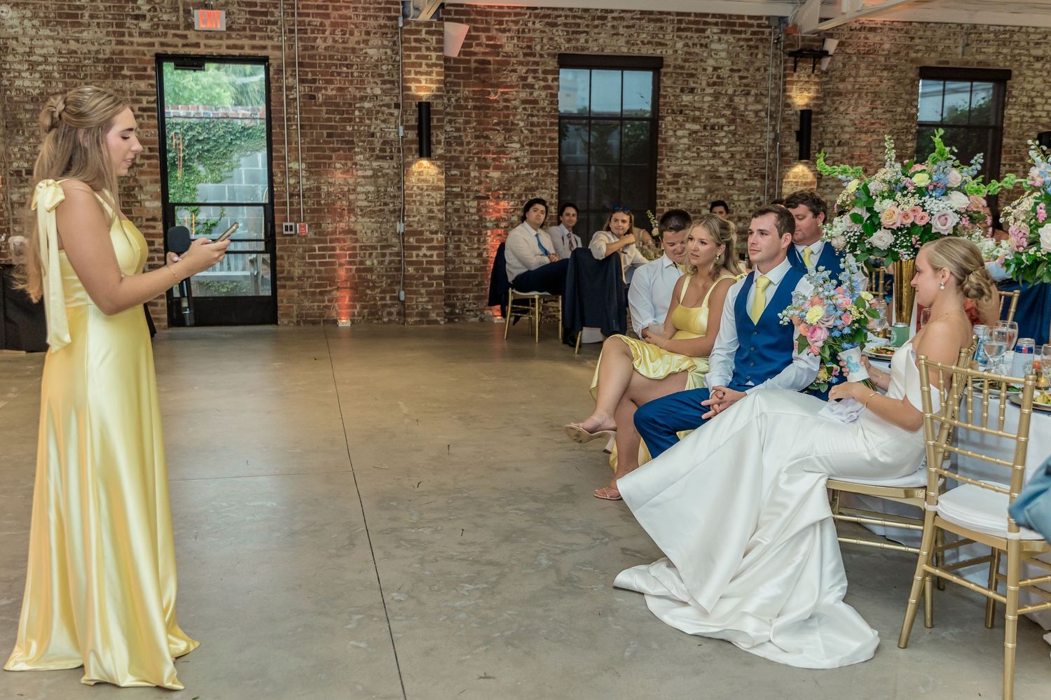 Elegant wedding reception setup in a rustic brick venue with gold chairs and white tablecloths.