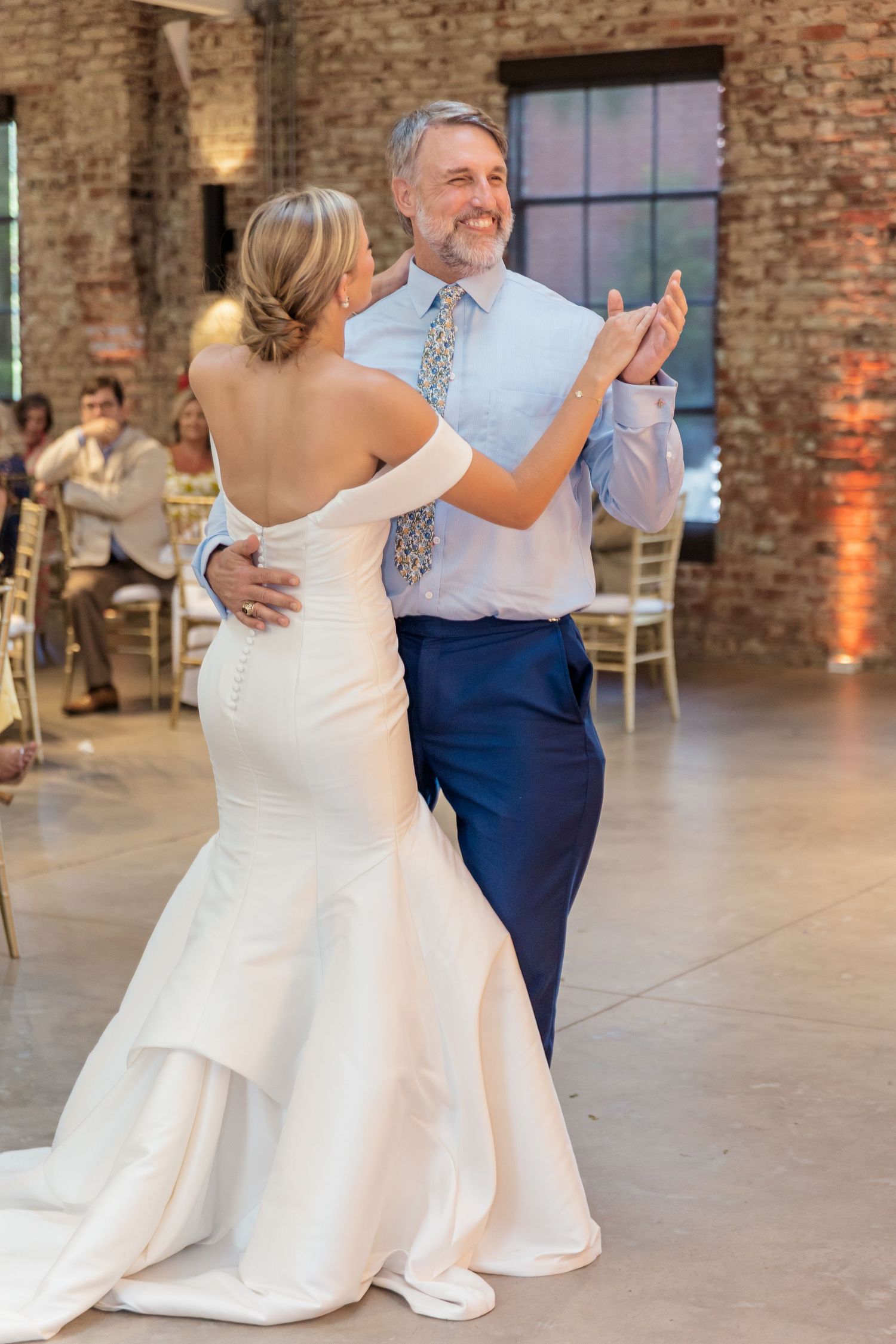 A couple shares their first dance at a rustic brick venue wedding reception, with guests seated at tables in the background.