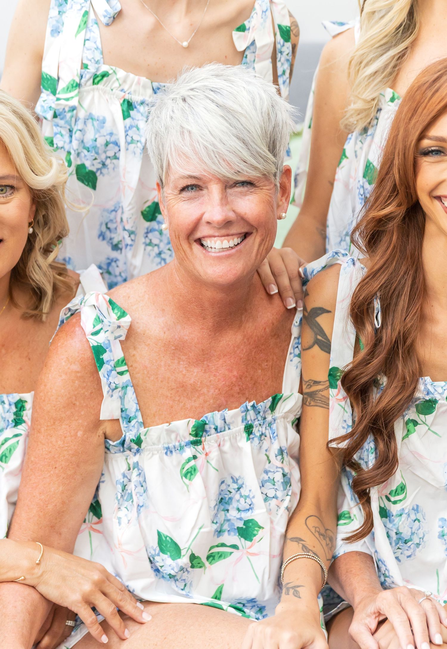 A group of women wearing matching floral print dresses in white and blue with green leaves smile together.