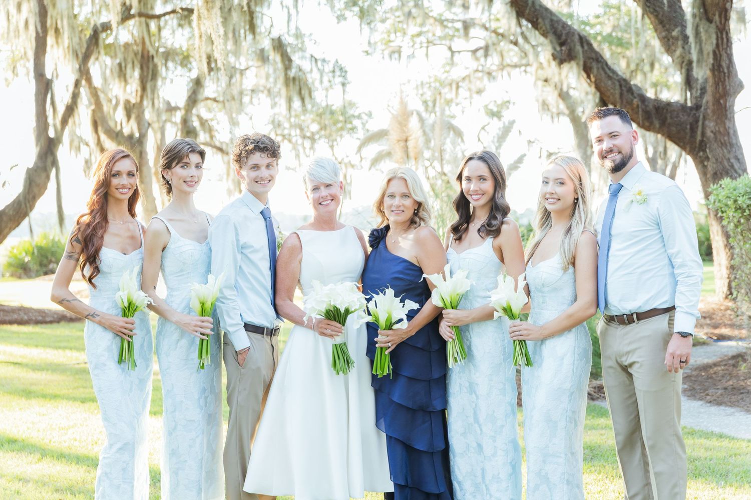 Wedding party in light blue and white attire poses under Spanish moss draped trees in an outdoor garden setting.