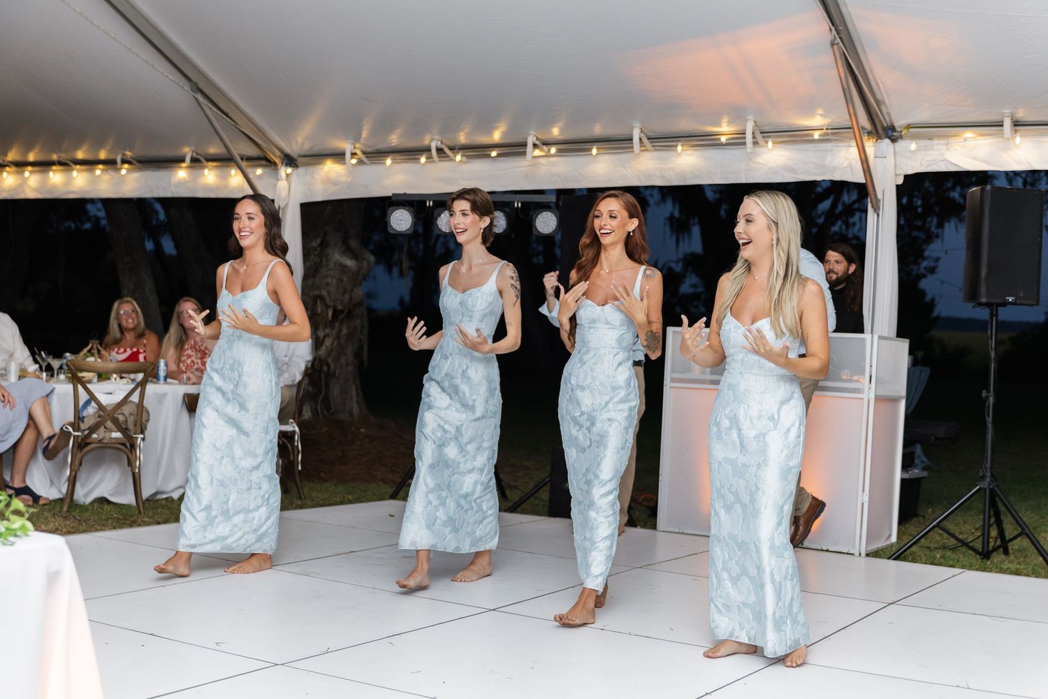 Bridesmaids in light blue dresses dance together on a white dance floor at an outdoor reception.