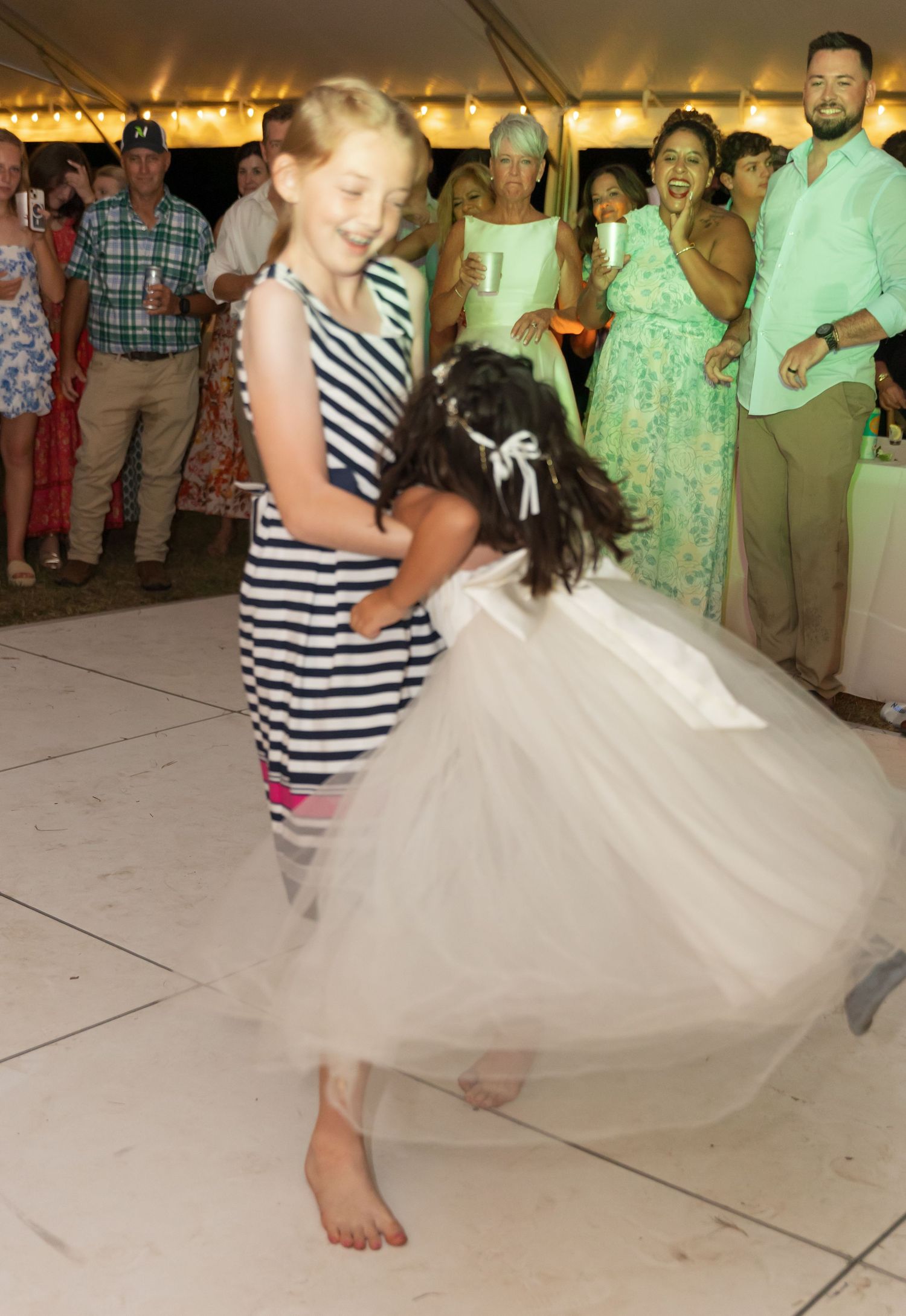 Two people dance together at a wedding reception under a tent, with one wearing a striped dress and the other in a white gown.