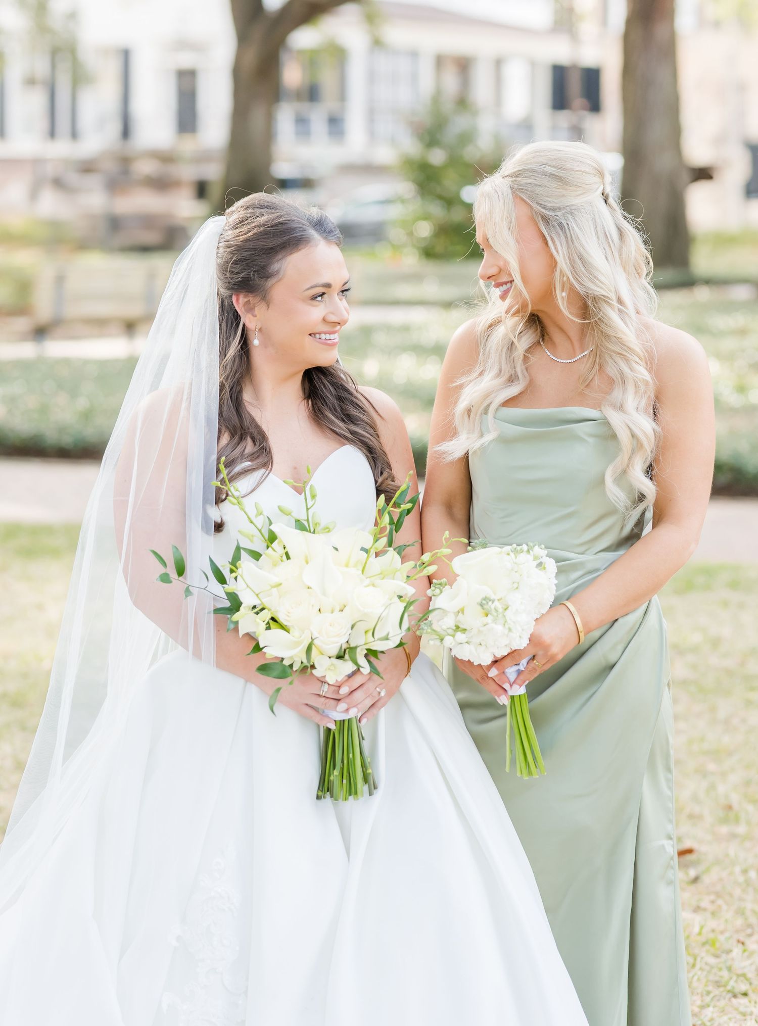 Wedding party members in sage green dresses and white gowns pose with bouquets in natural sunlight.