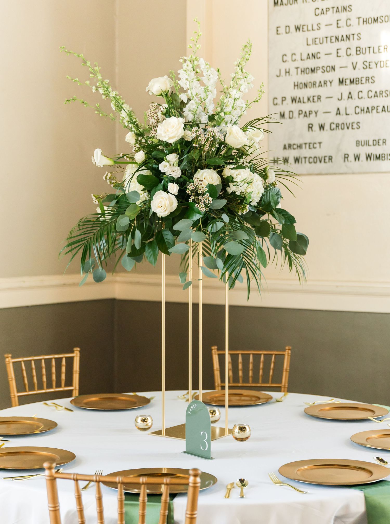 Wedding reception setup with gold chiavari chairs and tall white floral centerpieces.