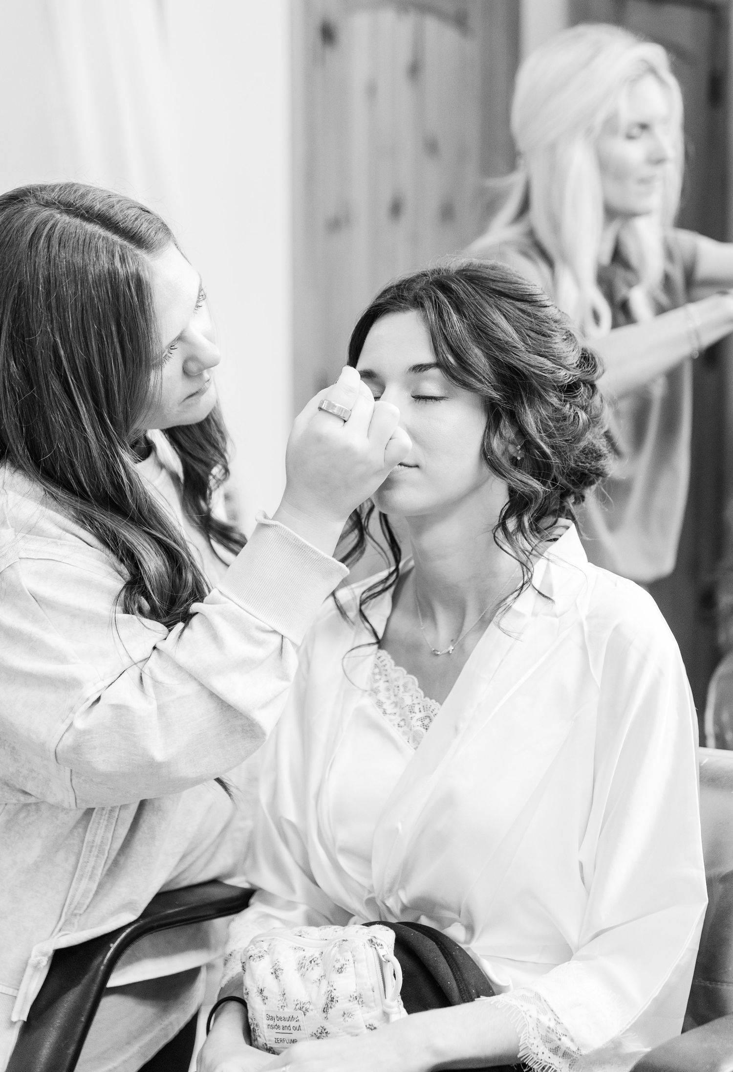 A makeup artist applies cosmetics to a bride getting ready in a white robe during wedding preparations.