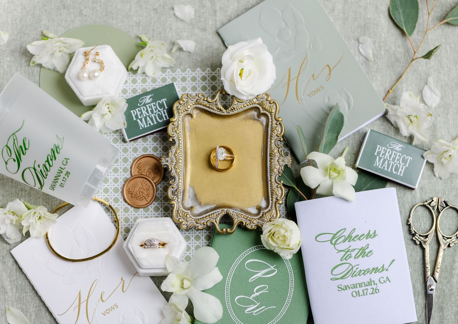 Elegant wedding details flatlay with white flowers, mint green stationery, and ornate gold frame on light background.