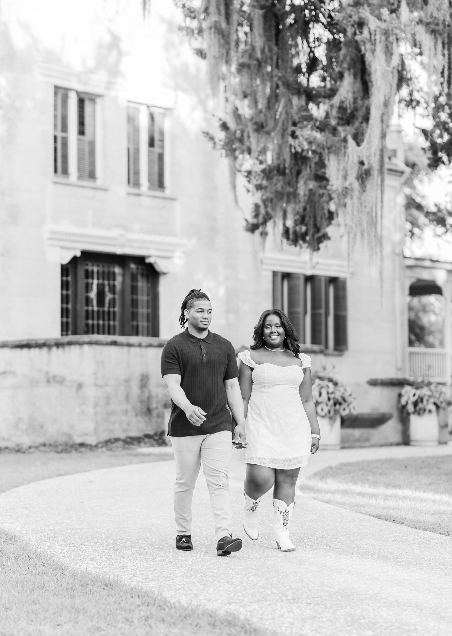Black and white engagement photo of couple walking along historic building grounds.