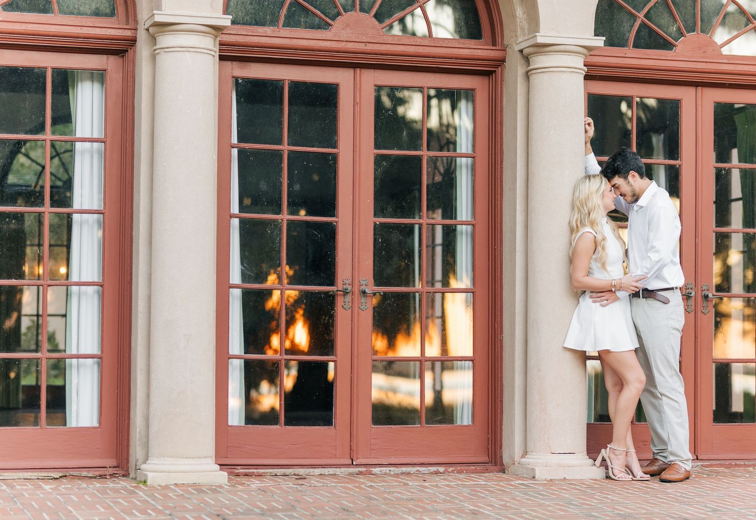 A couple in light summer attire shares a romantic moment between elegant columns and glass doors at sunset.