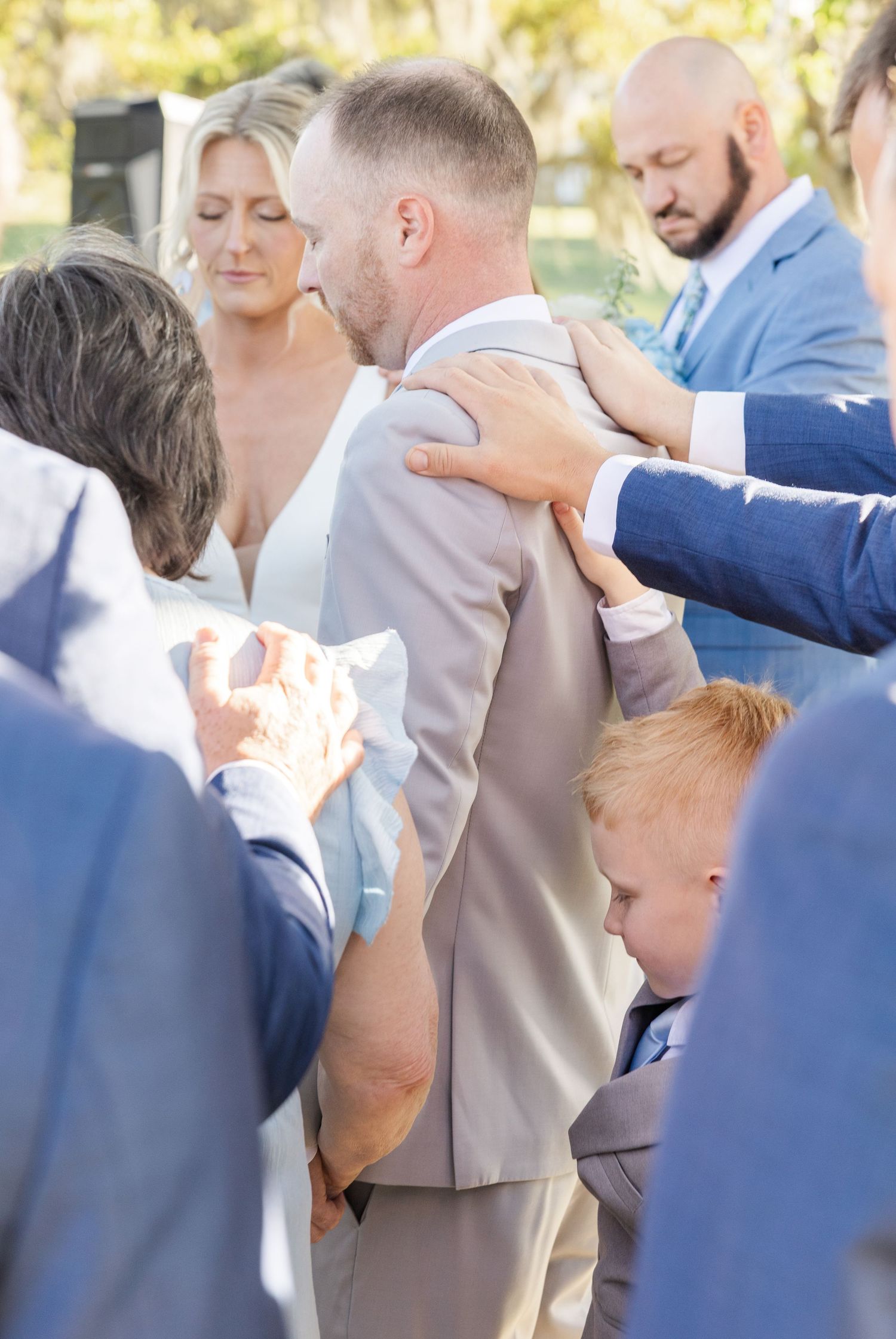 Wedding guests gather closely around the bride and groom in a group prayer during the outdoor ceremony.