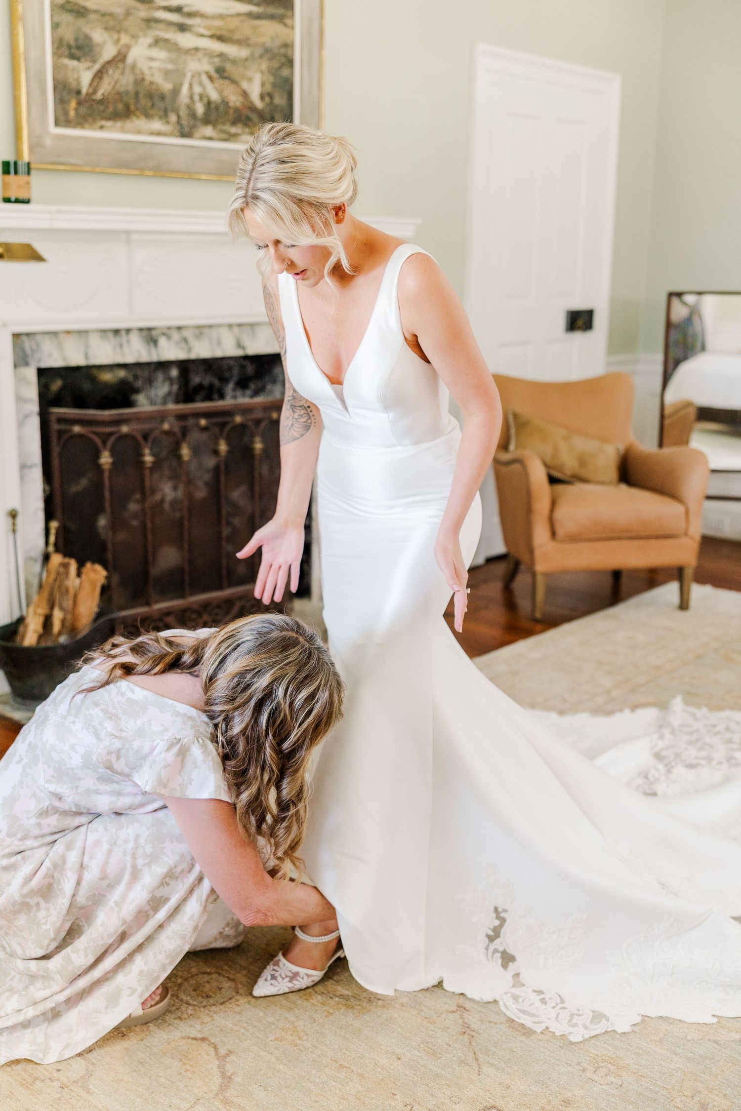 Bridesmaid kneels to adjust bride's shoes as she stands in elegant white gown before a fireplace.