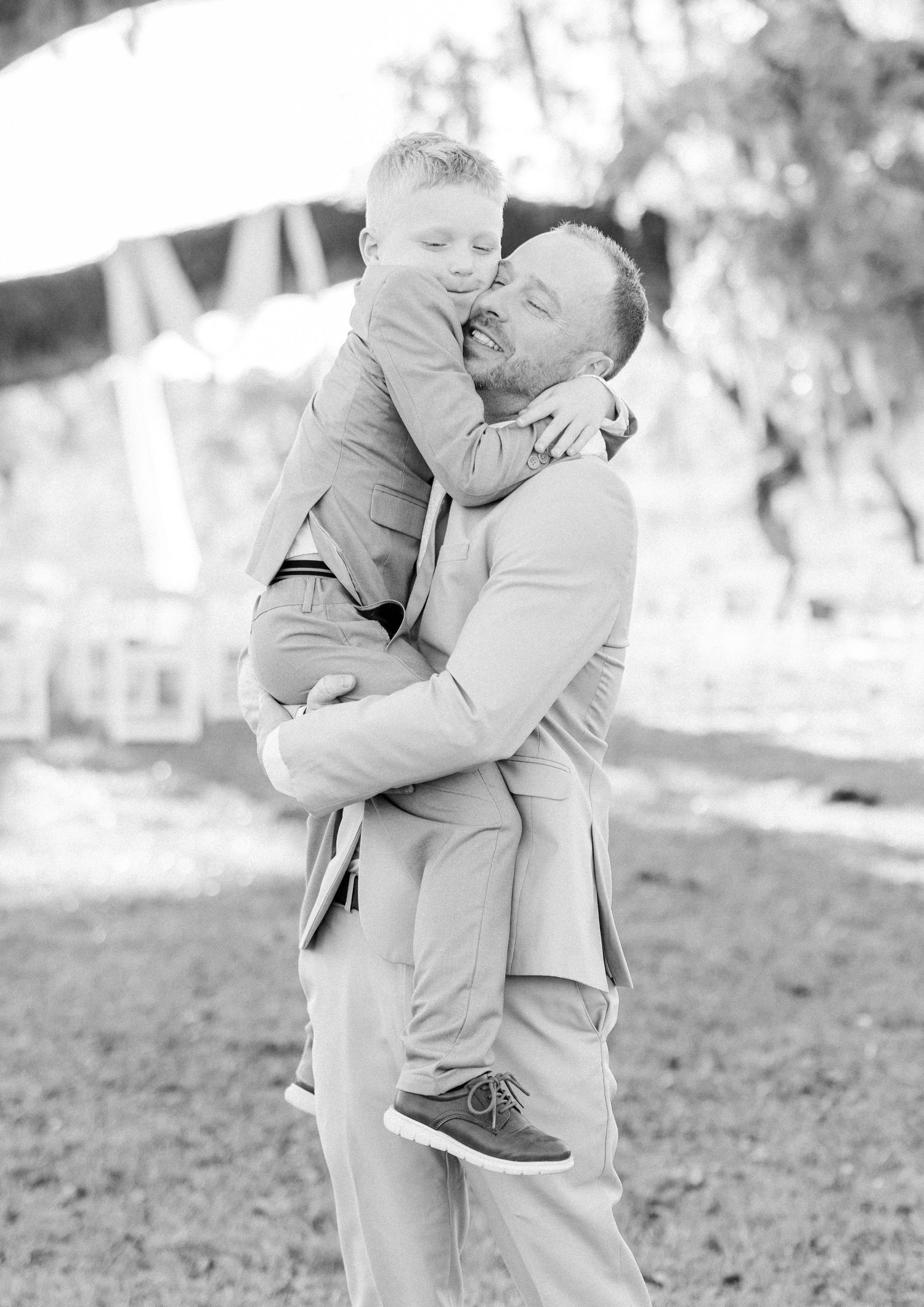 Black and white photo of groom holding young boy sharing a sweet kiss.