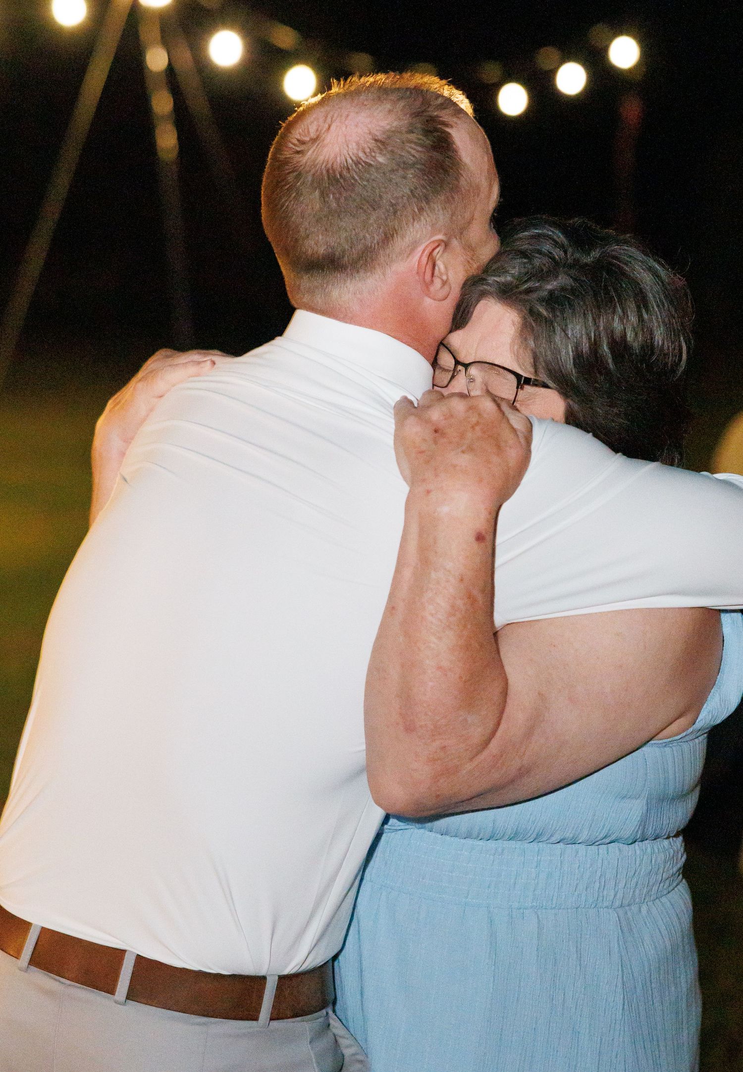 Groom in white shirt shares emotional dance embrace with older woman under string lights at night reception.