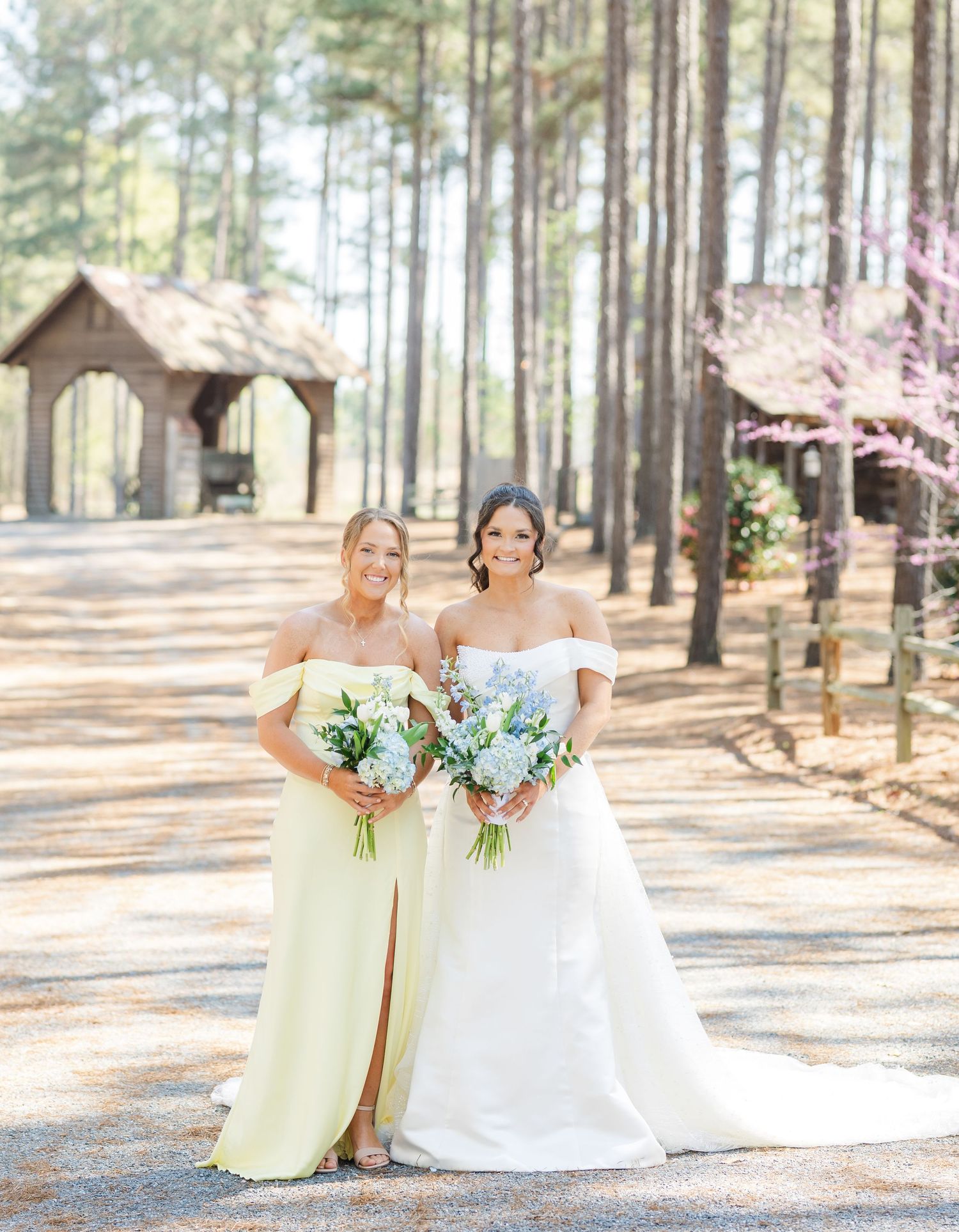 Bride in white off-shoulder gown and bridesmaid in yellow dress holding bouquets outdoors among pine trees.